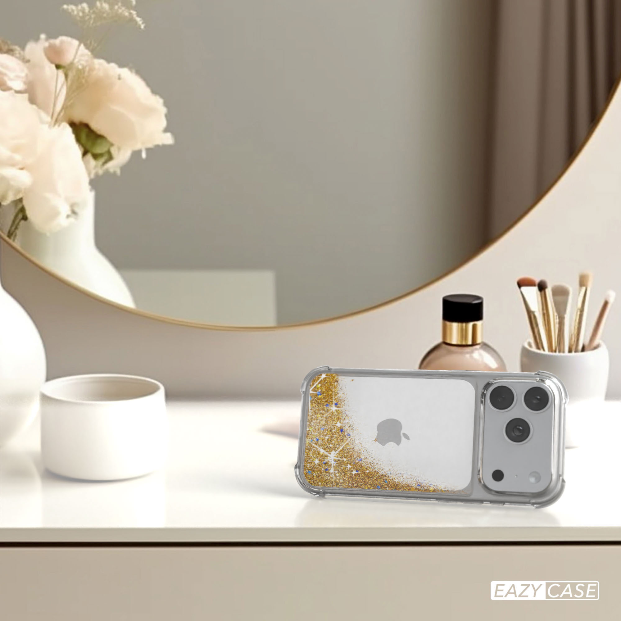 Ein iPhone in einer Glitzerhülle auf einem weißen Tisch. Blumen, Spiegel, Parfüm und Make-up-Pinsel im Hintergrund.
