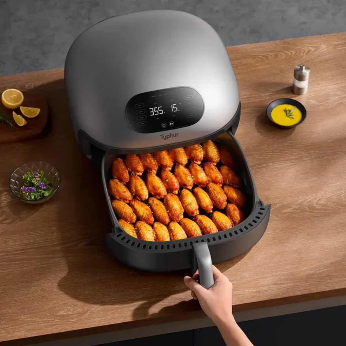 Ein Airfryer mit gekochten Chicken Wings. Zitronenscheiben, eine Salatschüssel und Sauce stehen auf der Holztheke.
