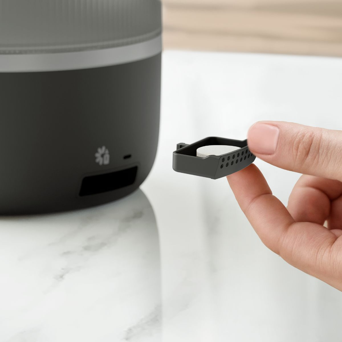 Ręka trzyma czarną tackę z białą tabletką. Czarno-szare urządzenie w tle na marmurowej powierzchni.