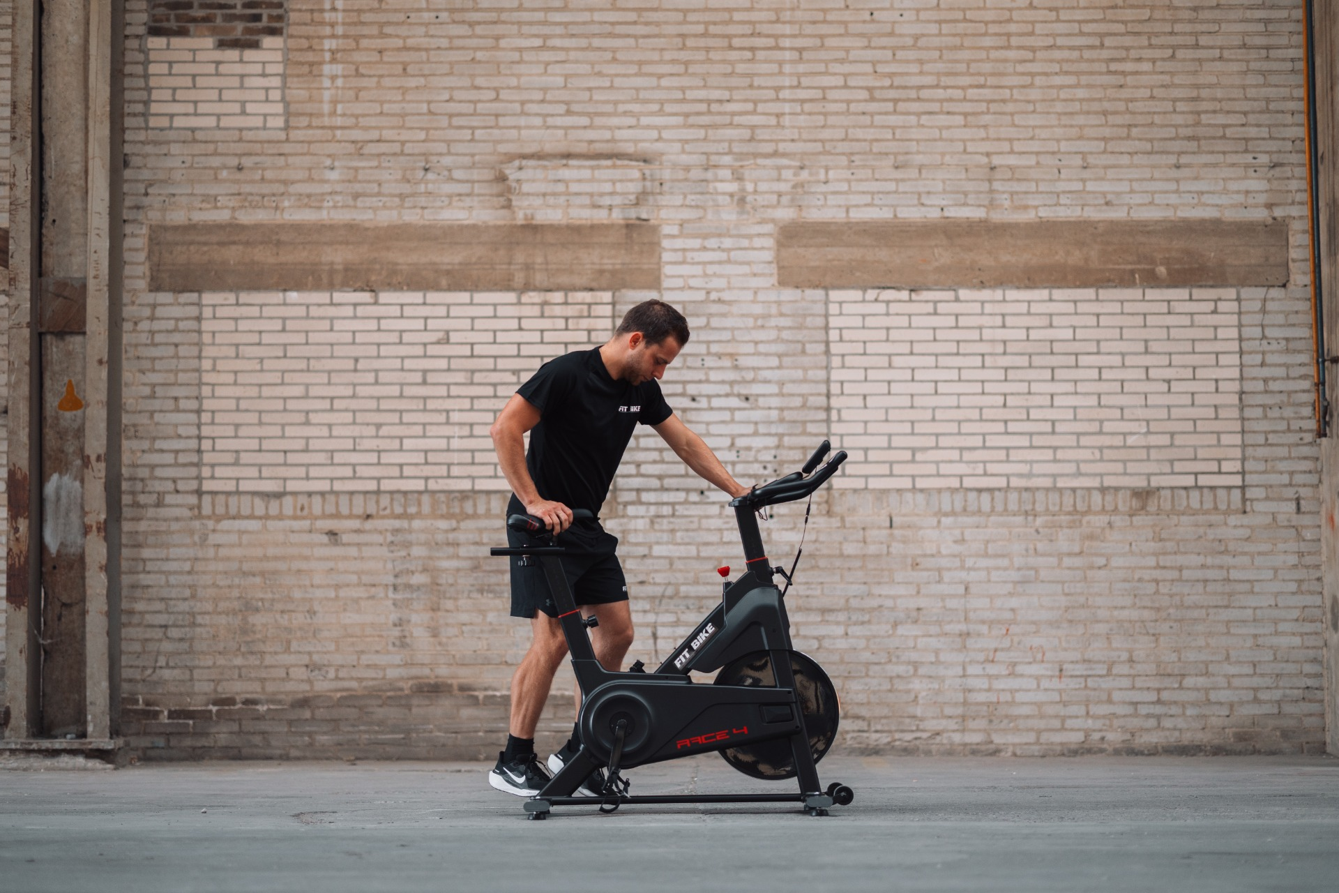Homme en tenue de sport noire près d'un vélo stationnaire devant un mur de briques. Il touche le vélo.