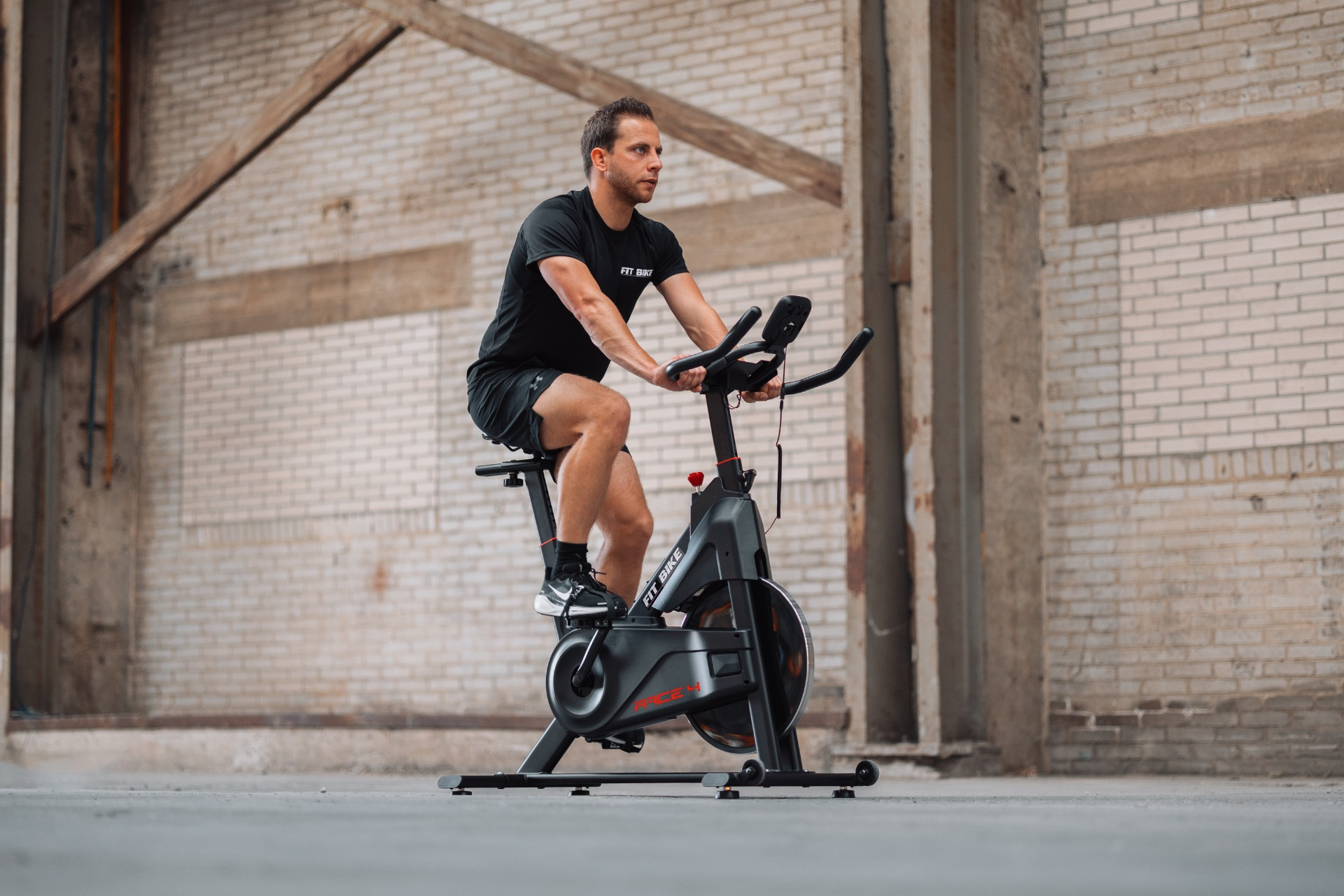 Un homme en tenue noire fait du vélo sur un vélo d'appartement. Le fond montre un mur de briques et un cadre industriel.