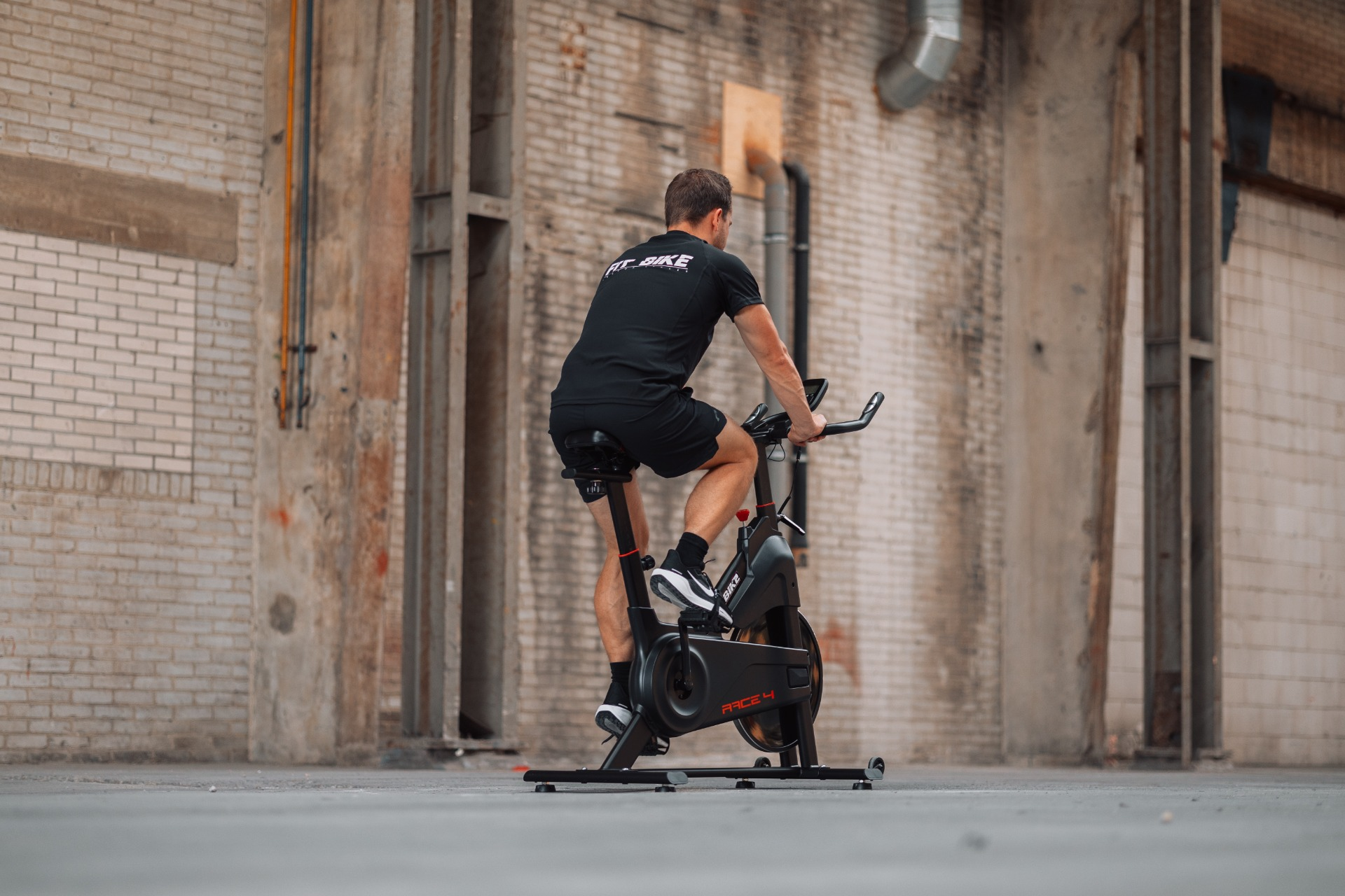Un homme sur un vélo d'appartement, dans un espace aux murs de briques. Il porte du noir et fait du vélo. Le vélo est noir et dit Race 4.