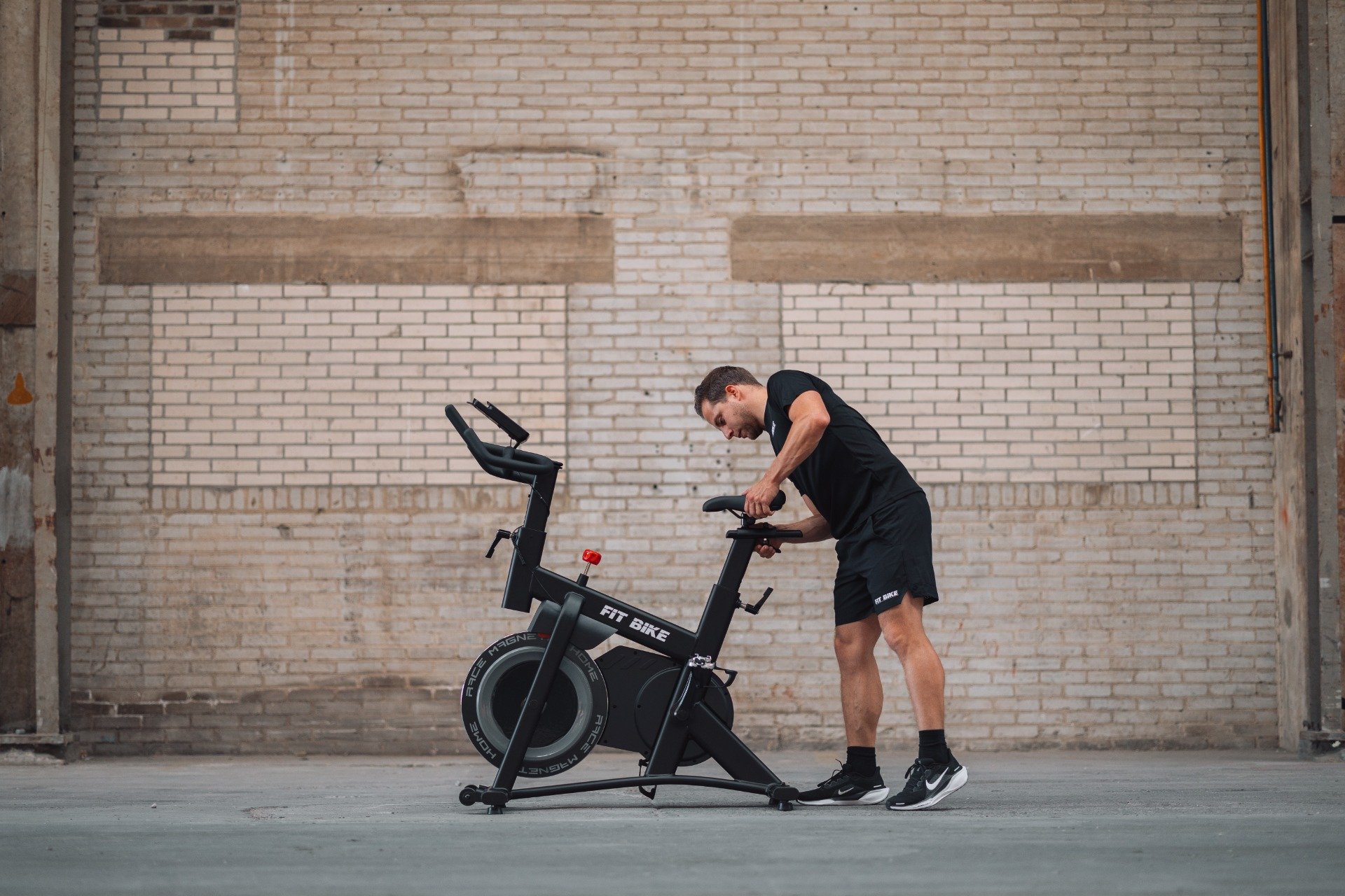 Un homme en noir ajuste un vélo stationnaire. Le vélo est noir avec 'FIT BIKE' dessus. Mur de briques en arrière-plan.
