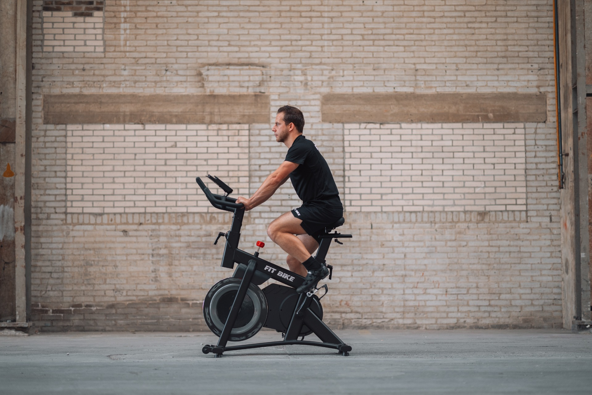 Un homme en vêtements noirs fait du vélo sur un vélo stationnaire noir. Un mur de briques est en arrière-plan.