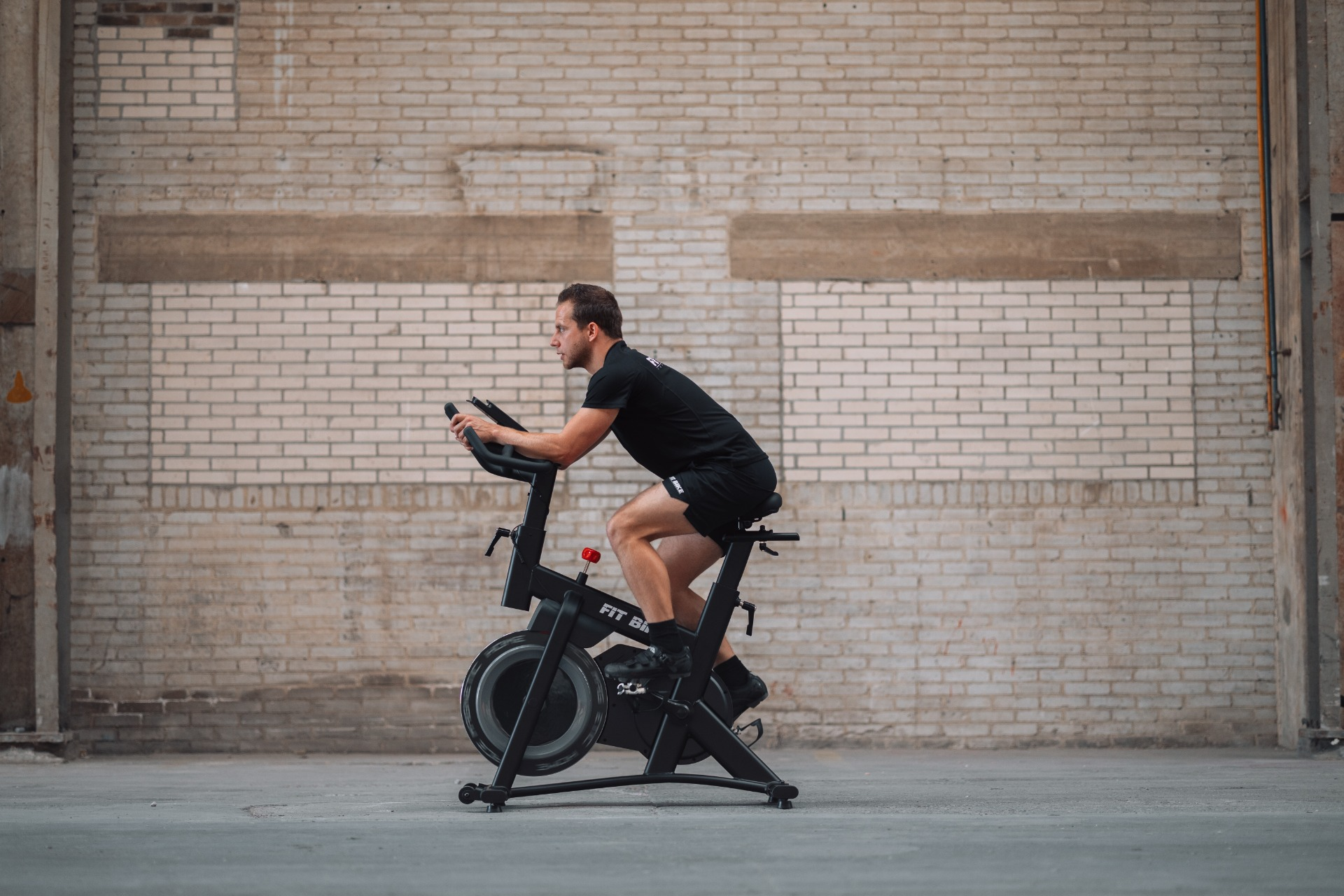 Un homme en vêtements noirs fait du vélo d'intérieur. Derrière lui se trouve un mur de briques. Le vélo dit 'Fit Bike'.