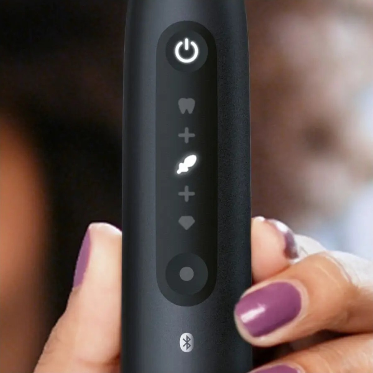 Ręka trzyma czarną elektryczną szczoteczkę do zębów. Szczoteczka ma podświetlany przycisk zasilania i wskaźniki trybu. Paznokcie są pomalowane na fioletowo.
