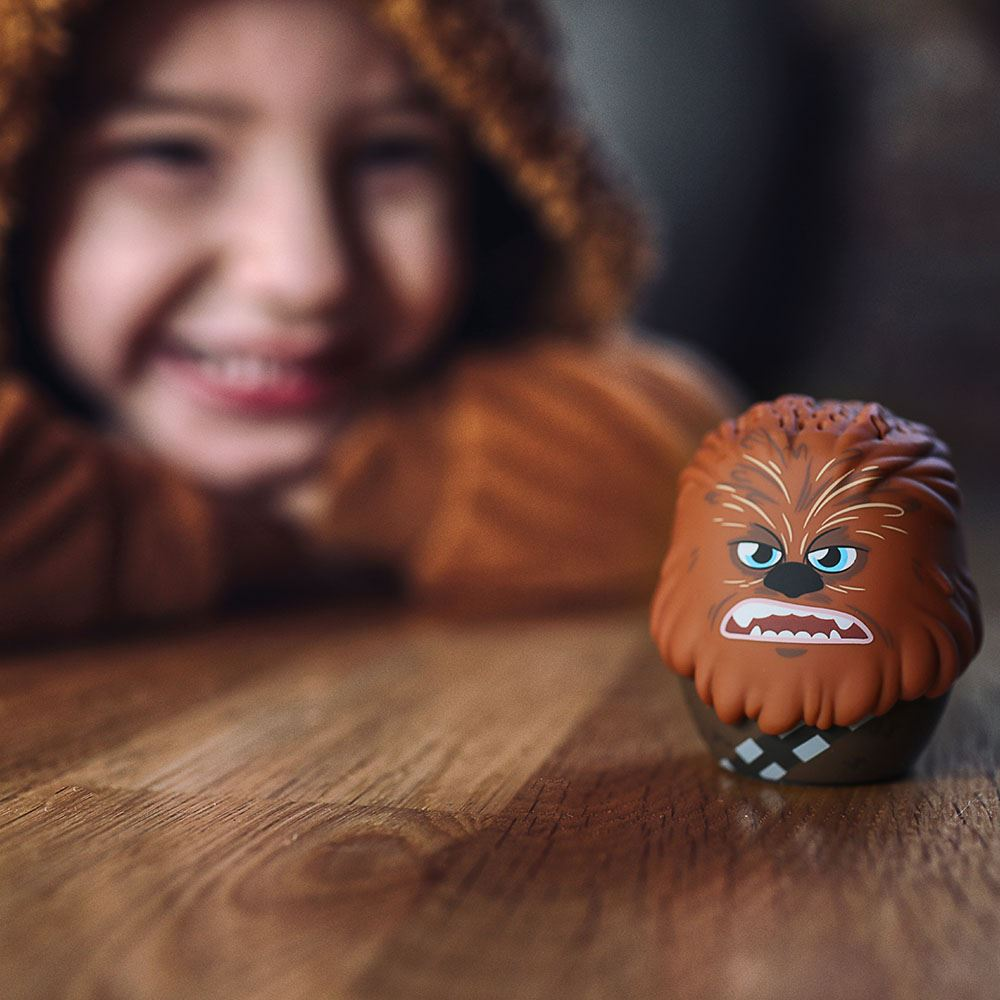 Kahverengi Chewbacca kostümü giymiş bir çocuk kameraya gülümsüyor. Ahşap bir masada Chewbacca oyuncağı duruyor.