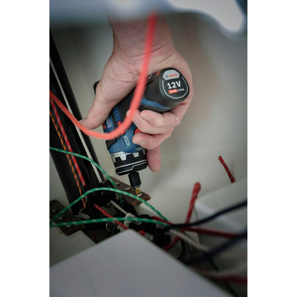 Dłoń trzyma wiertarkę Bosch 12V. W tle kable i liny. Wiertarka służy do naprawy czegoś na łodzi.