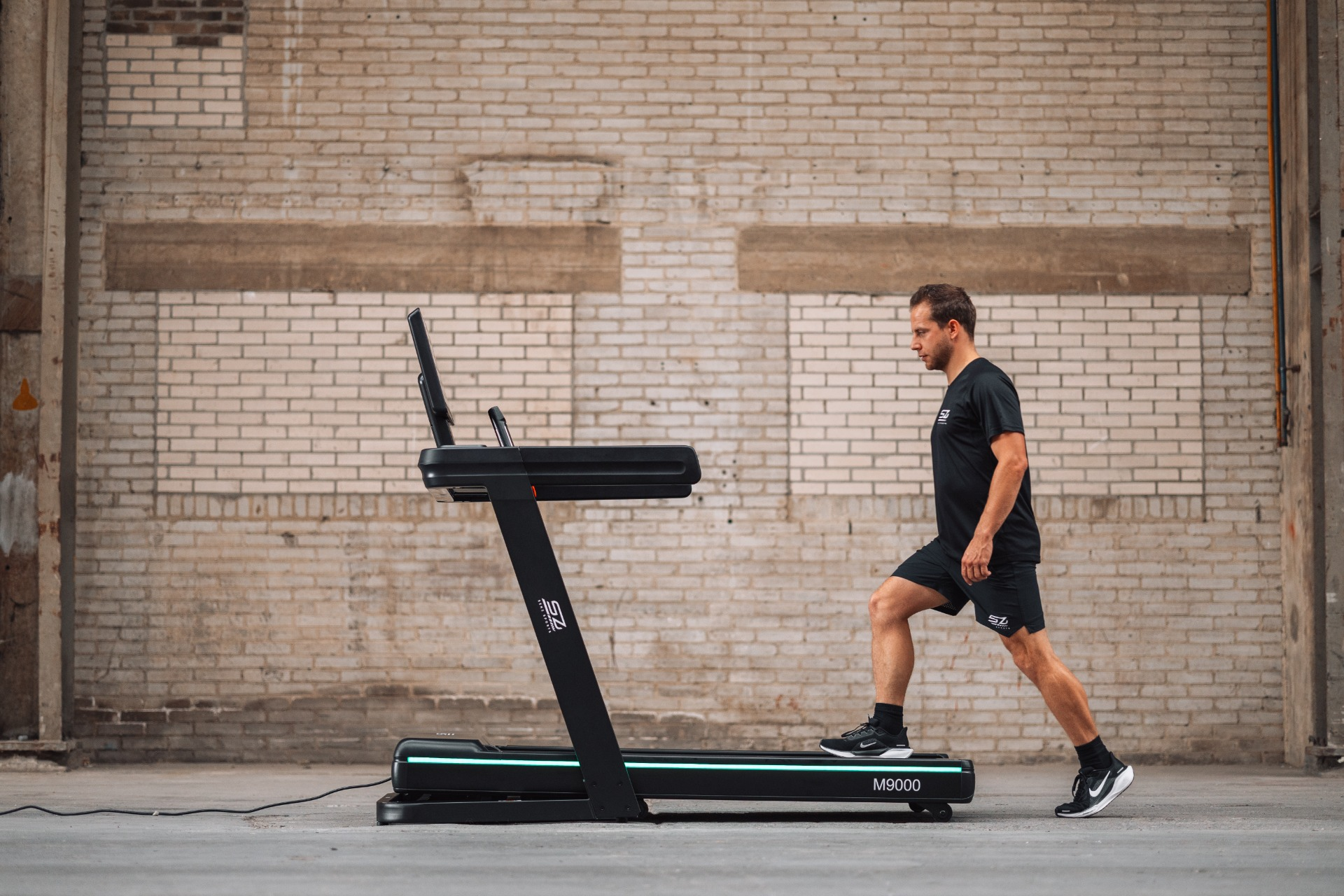 A man in black sportswear is running on a black treadmill. The background is a brick wall in a warehouse.