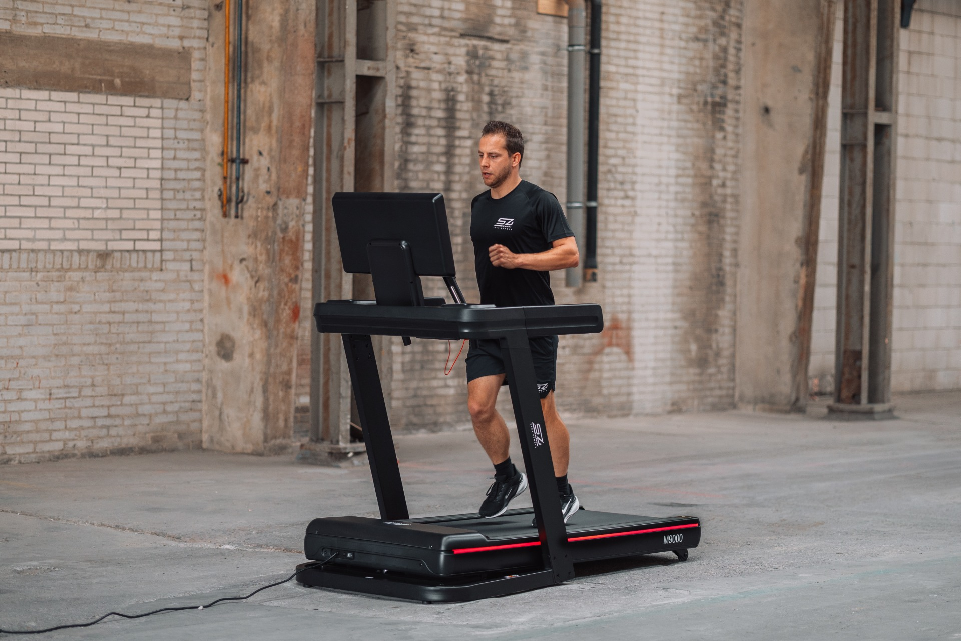 A man runs on a treadmill. He wears black athletic clothing. The treadmill is black with red accents. Background is brick.