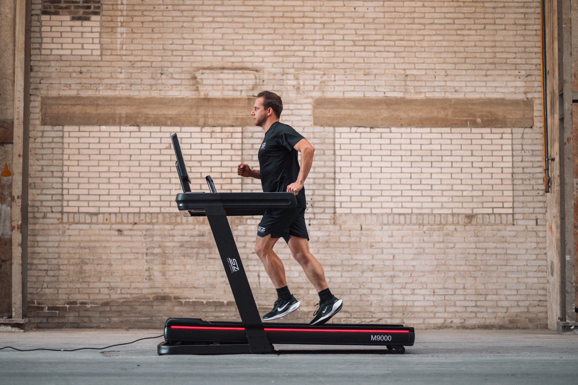 A man runs on a treadmill. He wears black sportswear. The background is a brick wall. The treadmill is black and red.