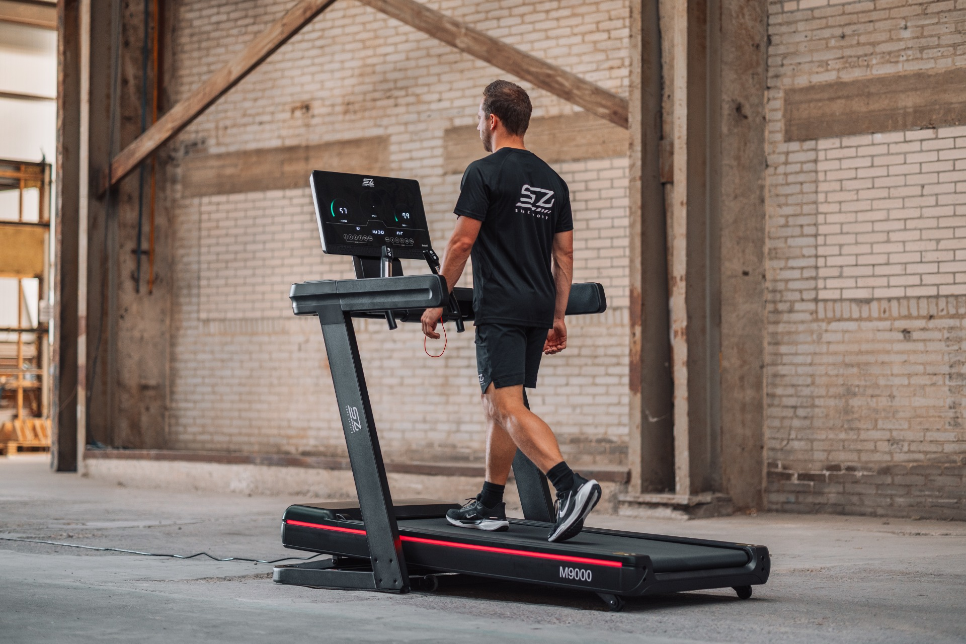 Man walking on a treadmill in a warehouse. He wears black shorts and a shirt. The treadmill has a screen.