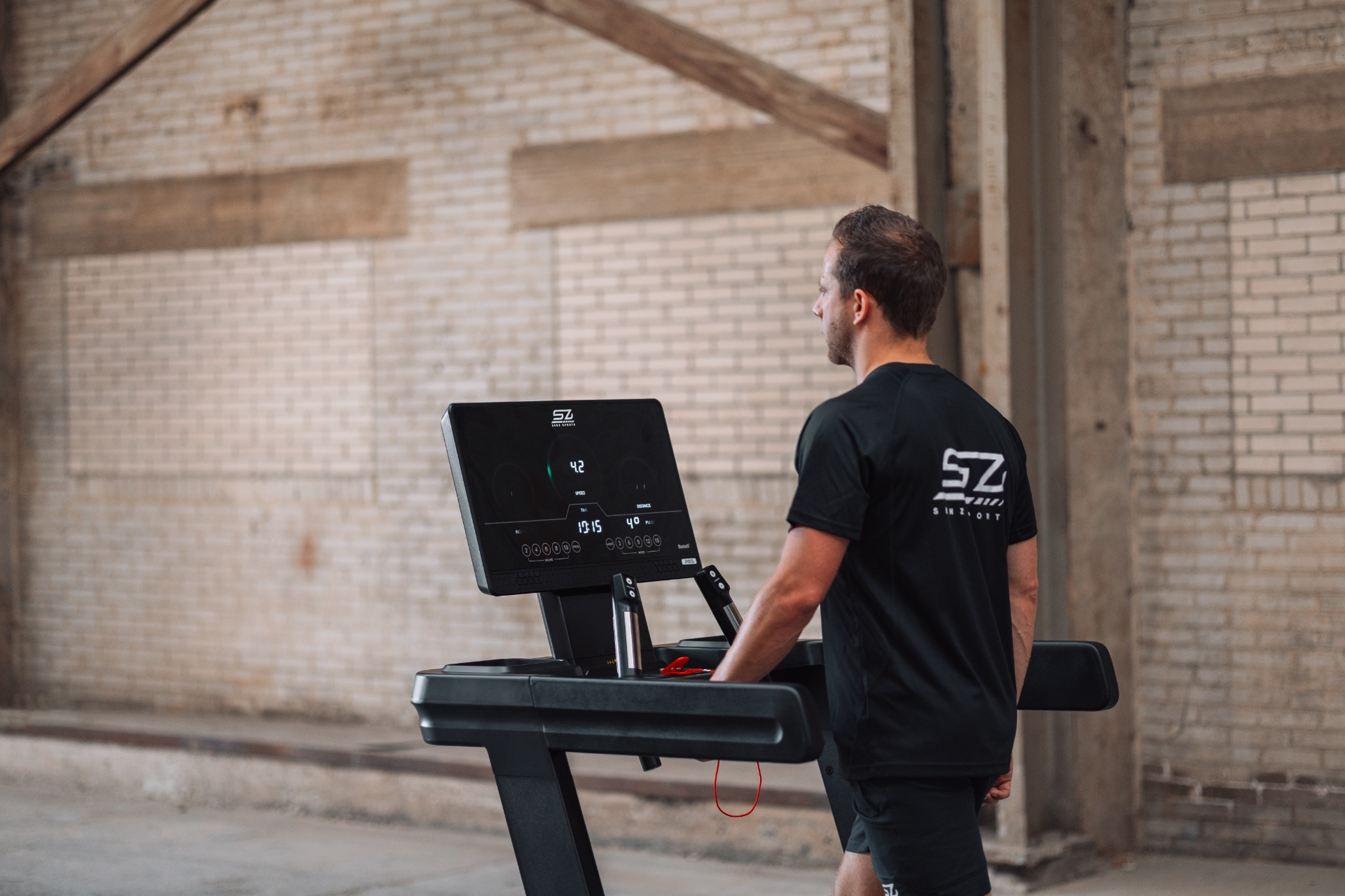Man stands by a treadmill in a brick building. The treadmill display shows speed and other metrics. He wears a black shirt.