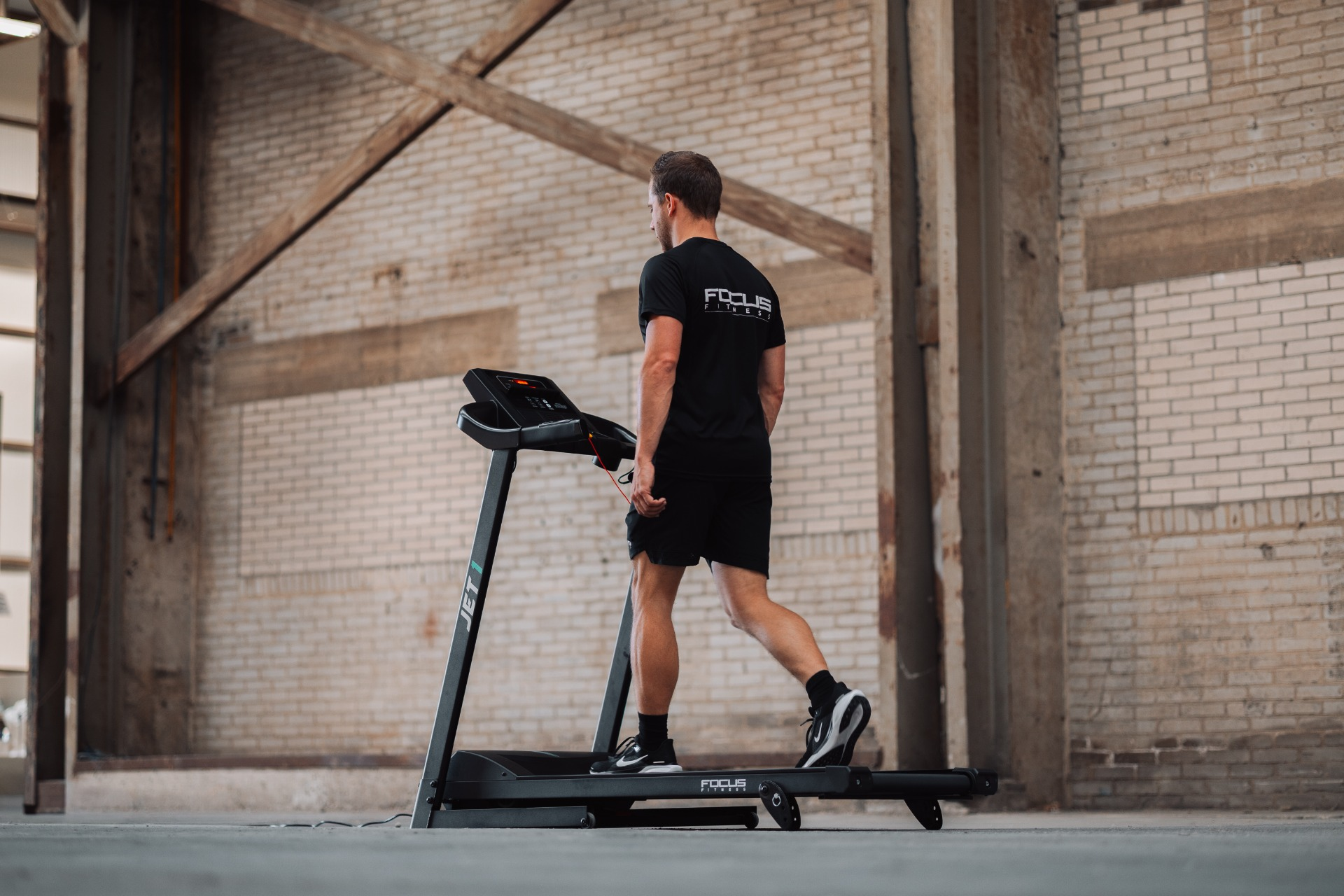 A man in black shorts and shirt runs on a treadmill. The background shows a brick wall and industrial structure.