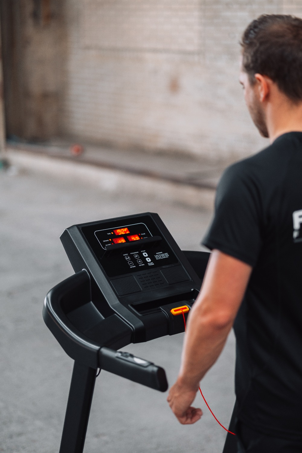 A man stands near a treadmill. The machine's display is lit. The background is a concrete wall.