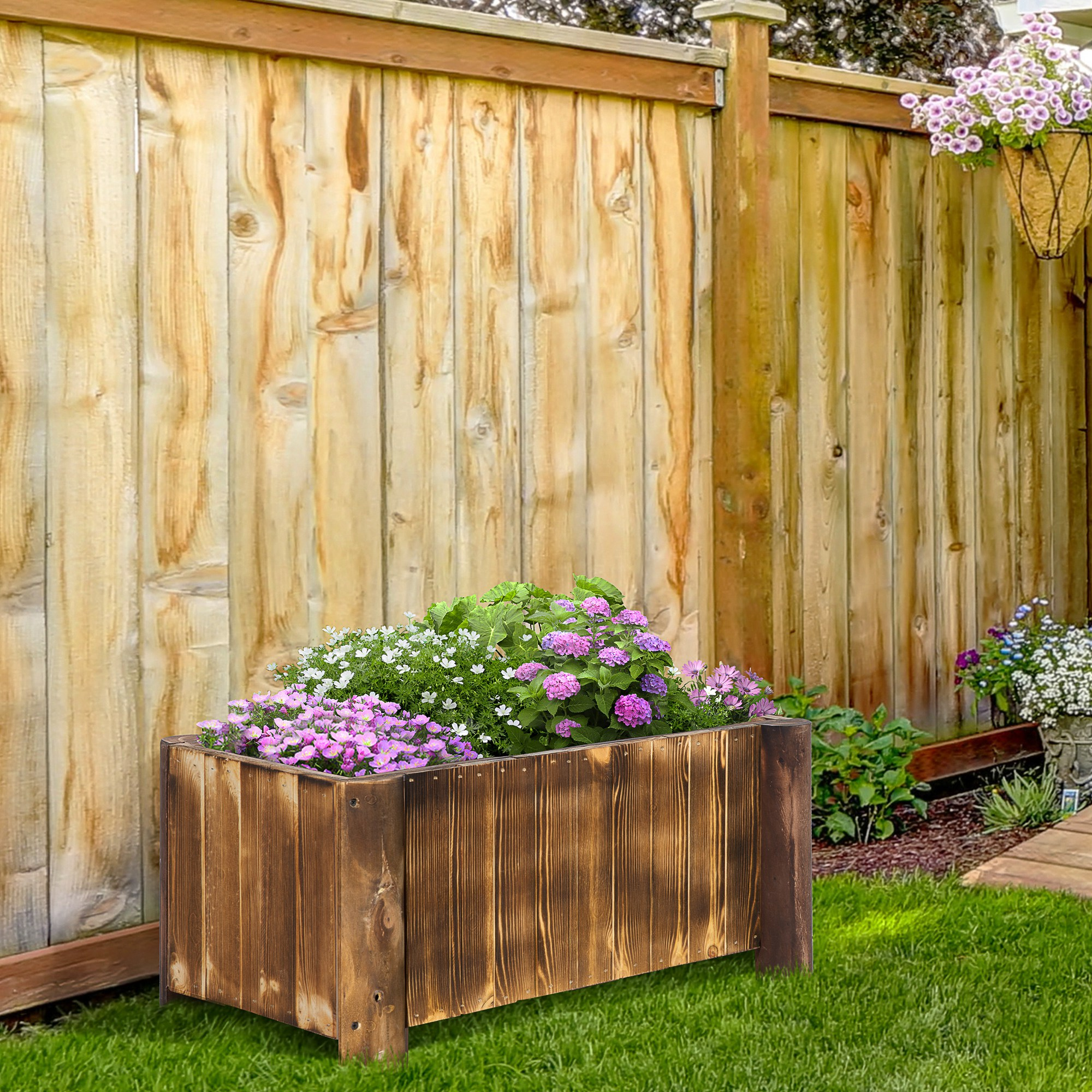 Fioriera in legno con fiori viola, rosa e bianchi sull'erba. Una staccionata in legno sullo sfondo.