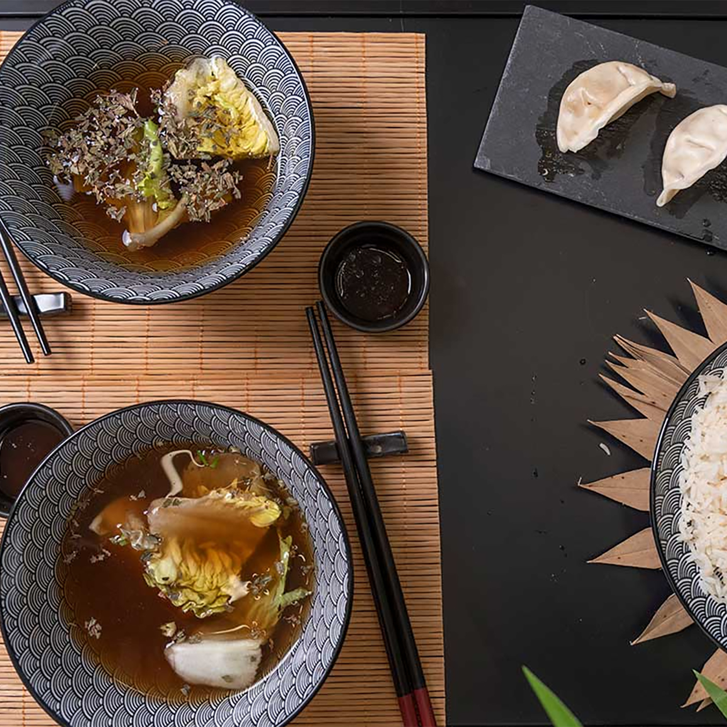 Vista dall'alto di piatti asiatici: ciotole di zuppa con cavolo, ravioli e riso. Bacchette e piccole ciotole di salsa su un tappetino di bambù.