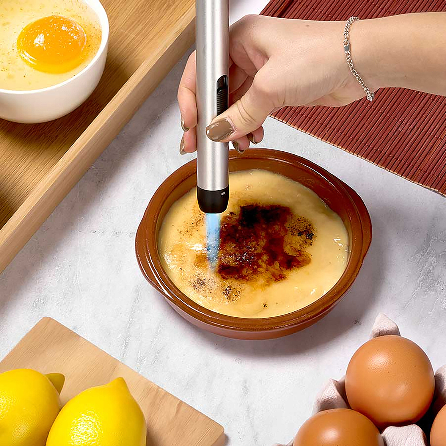Una mano usa un soplete para caramelizar crema catalana. También se ven huevos, limones y un cuenco con natillas.