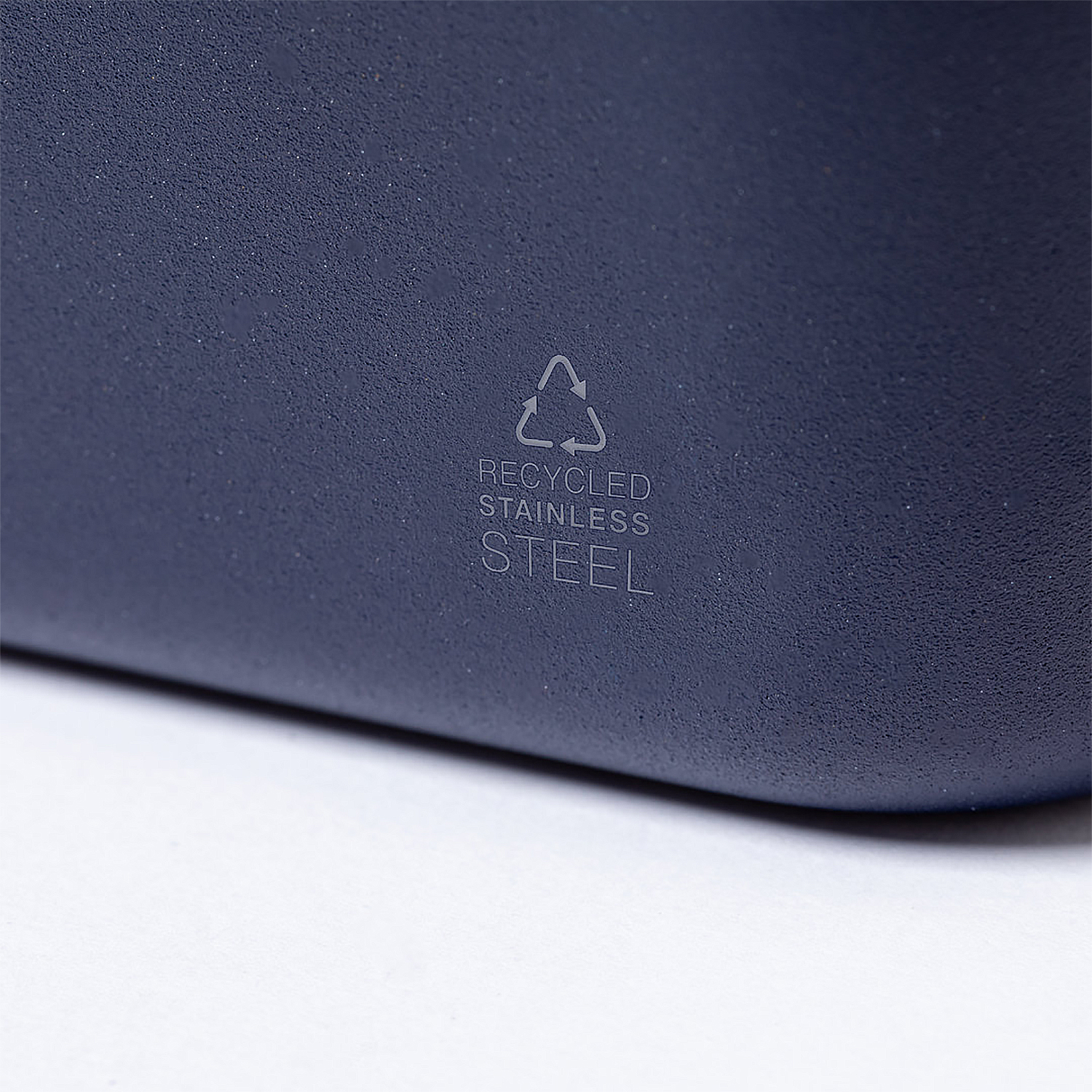 Nahaufnahme einer dunkelblauen, strukturierten Oberfläche mit einem Recycling-Symbol und den Worten 'Recycled Stainless Steel'.