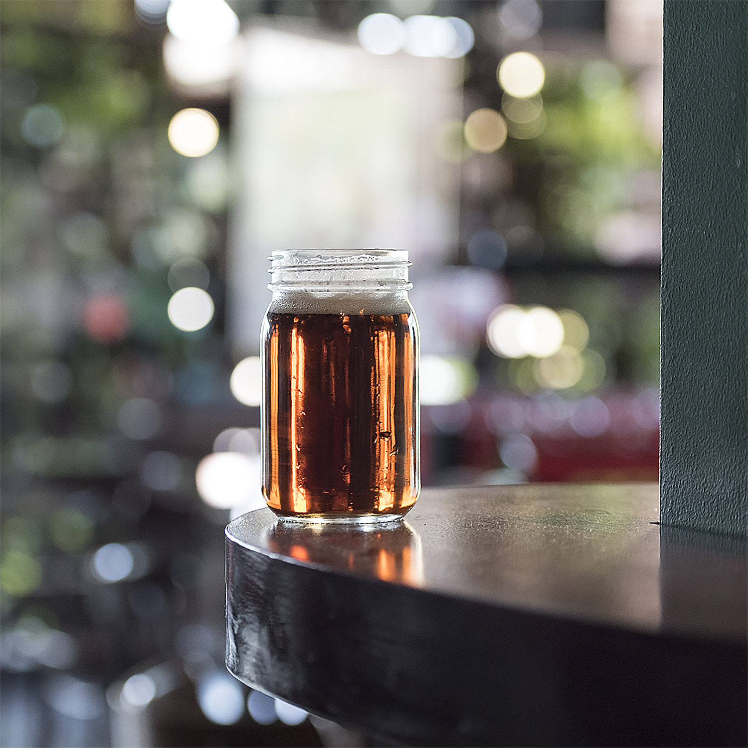 Un barattolo di vetro pieno di liquido ambrato, probabilmente birra, è su una superficie di legno scuro. Lo sfondo mostra un bar sfocato.