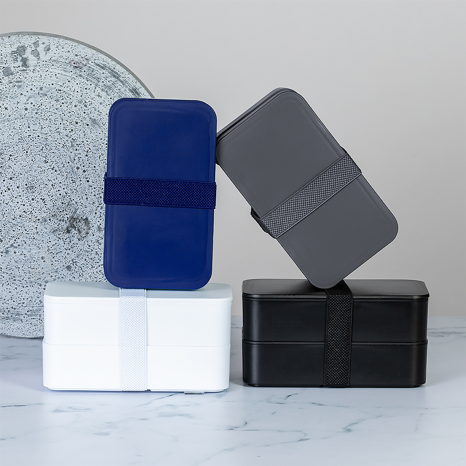 Cuatro fiambreras apiladas: azul, gris, blanco y negro. Cada una tiene una correa. Están sobre una superficie de mármol. Un plato texturizado en el fondo.