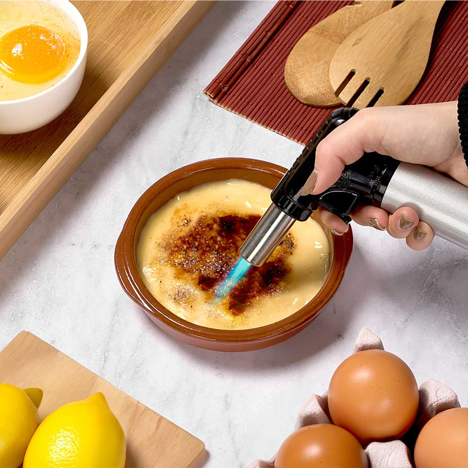 Una mano flambea crema catalana. Huevos, limones y utensilios de madera rodean el postre, sobre una superficie similar al mármol.