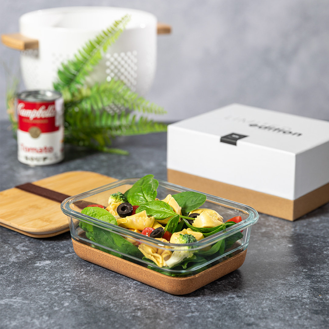Un recipiente de vidrio con ensalada está sobre una mesa. Una lata de sopa, una caja y un helecho están en el fondo. La ensalada tiene aceitunas y tomates.