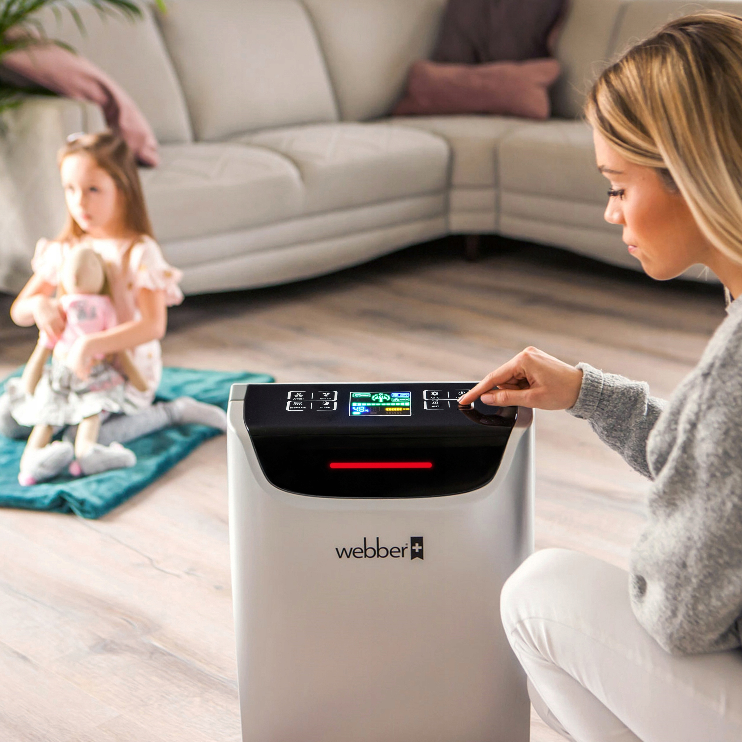 Kobieta obsługuje oczyszczacz powietrza, a dziecko z lalką siedzi na macie. W tle kanapa.