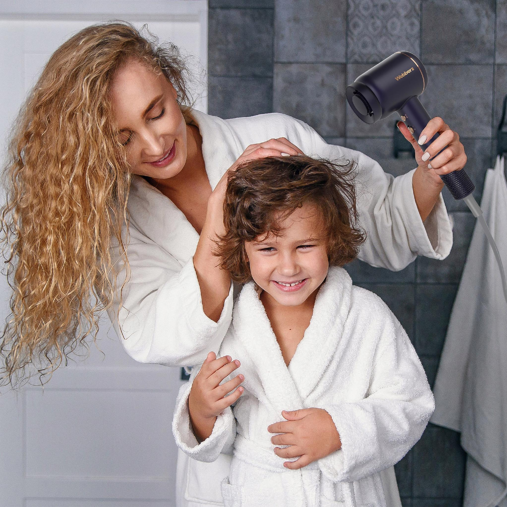 Kobieta suszy włosy dziecka suszarką. Oboje mają na sobie białe szlafroki. Dziecko się uśmiecha.