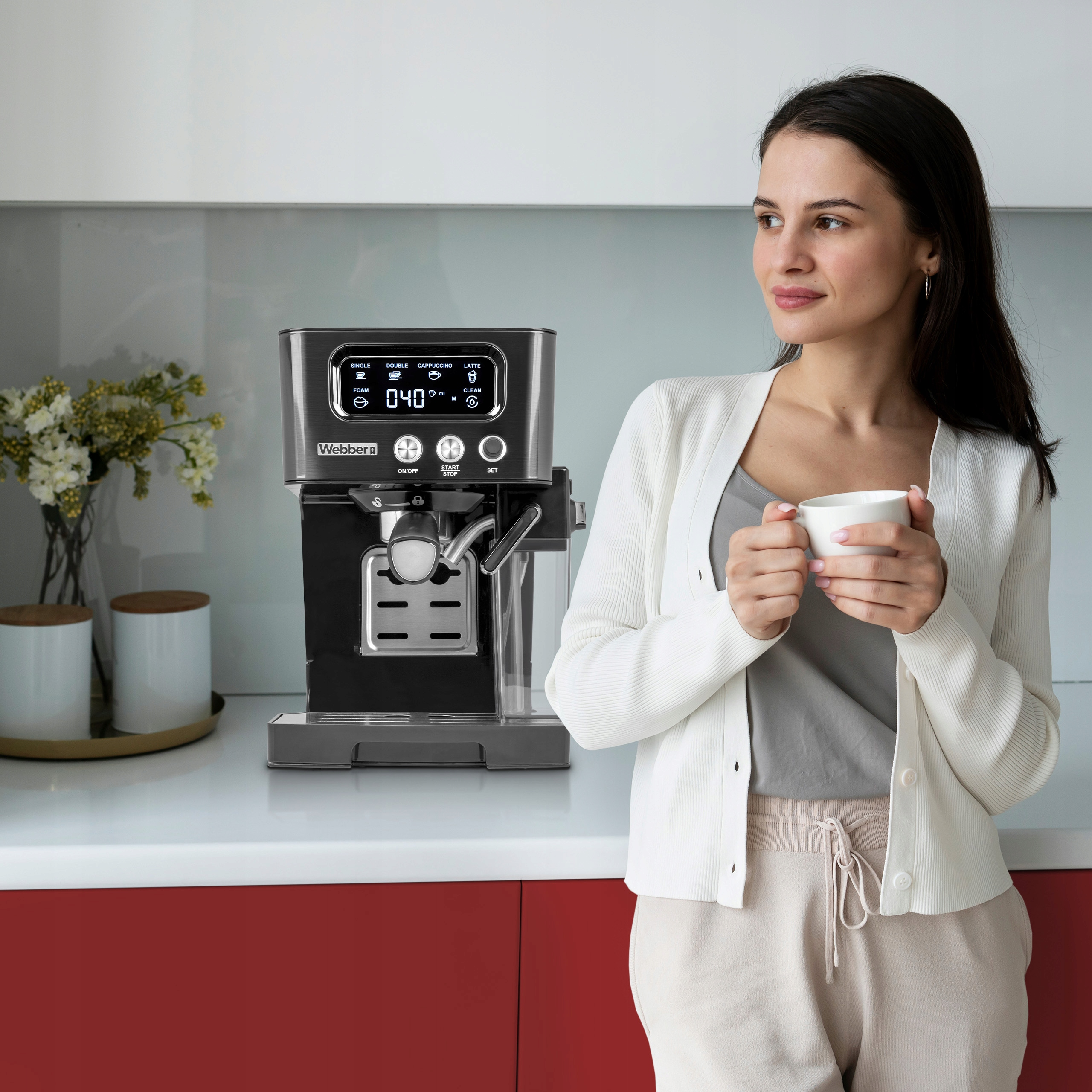 Kobieta trzyma biały kubek, stojąc blisko ekspresu do kawy. Maszyna ma cyfrowy wyświetlacz. Otoczenie kuchni.