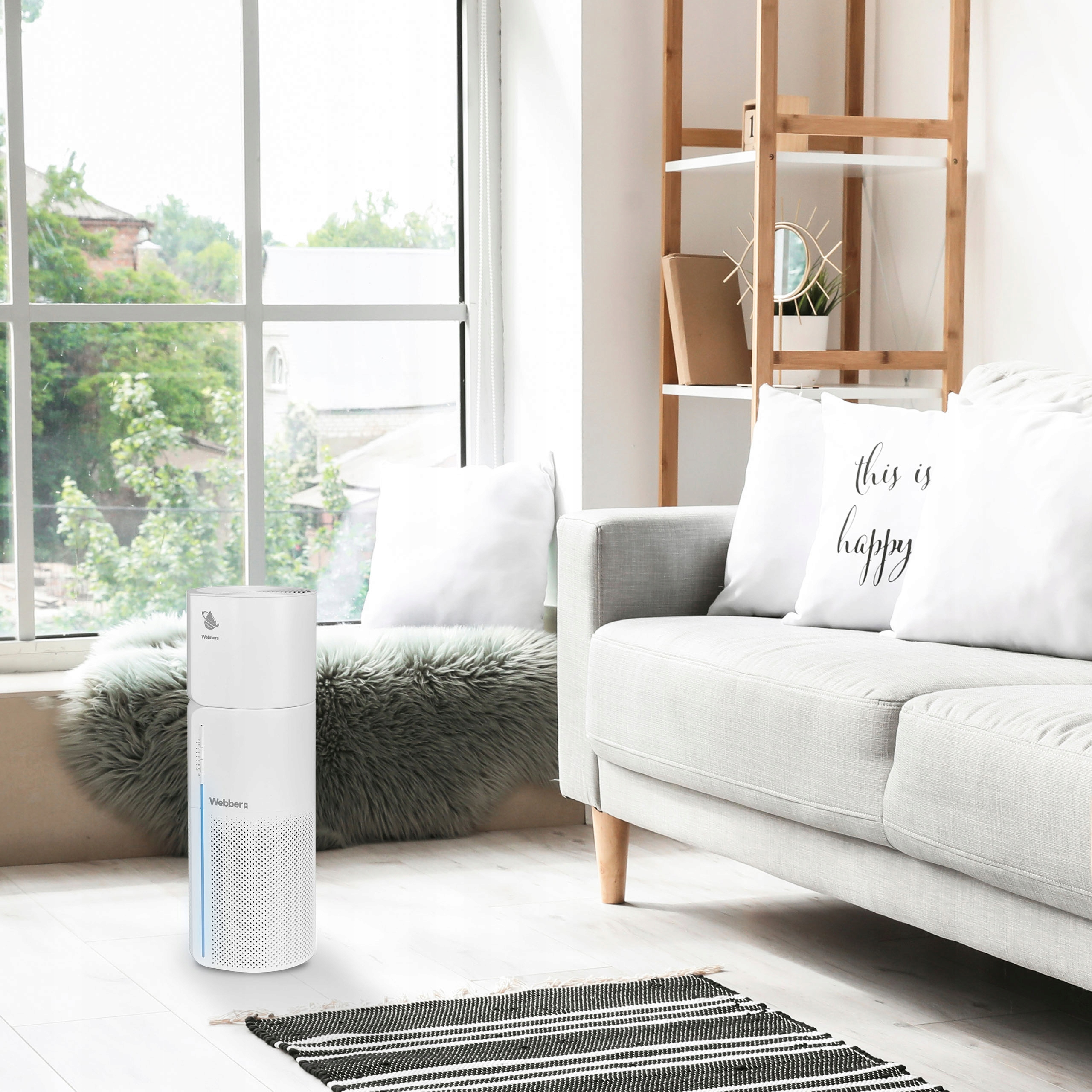 Biały oczyszczacz powietrza stoi blisko okna. Po prawej stronie szara kanapa z poduszkami. Na podłodze dywan.