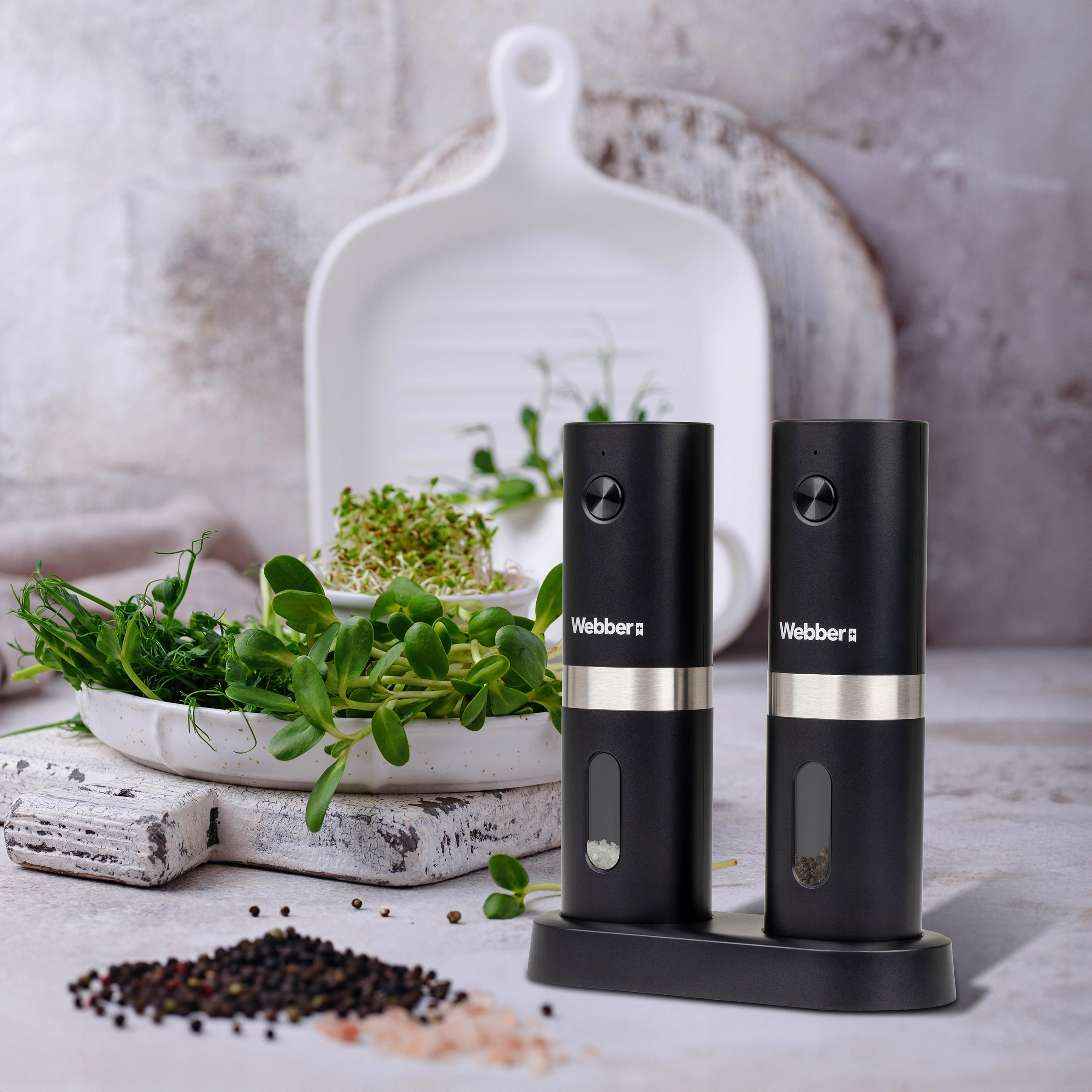 Dwa czarne młynki do soli i pieprzu z marką Webber, mikrozielenią, pieprzem i białymi deskami do krojenia.