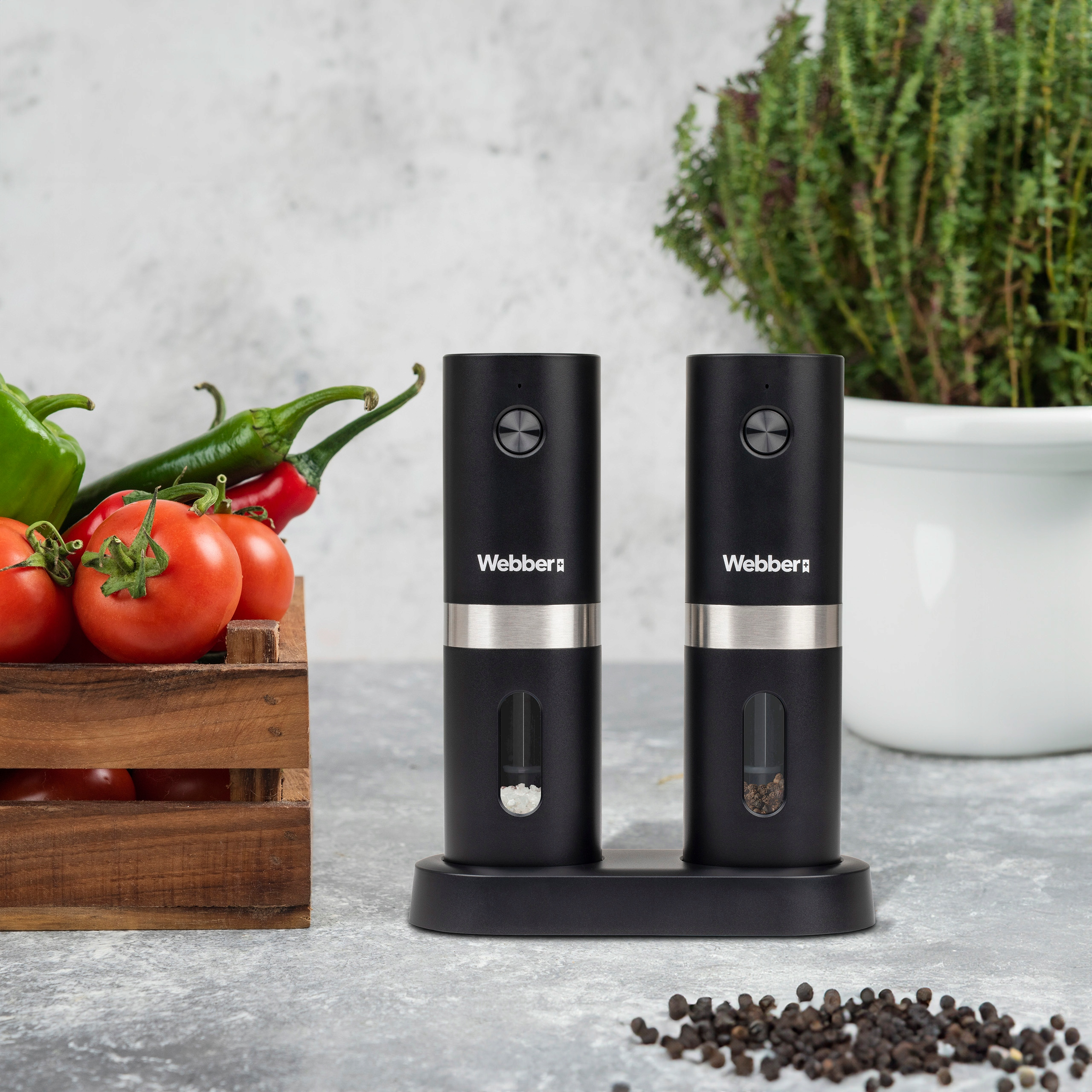Dwa czarne młynki do soli i pieprzu, pomidory, papryka i zioła na stole. Ziarna pieprzu rozsypane.