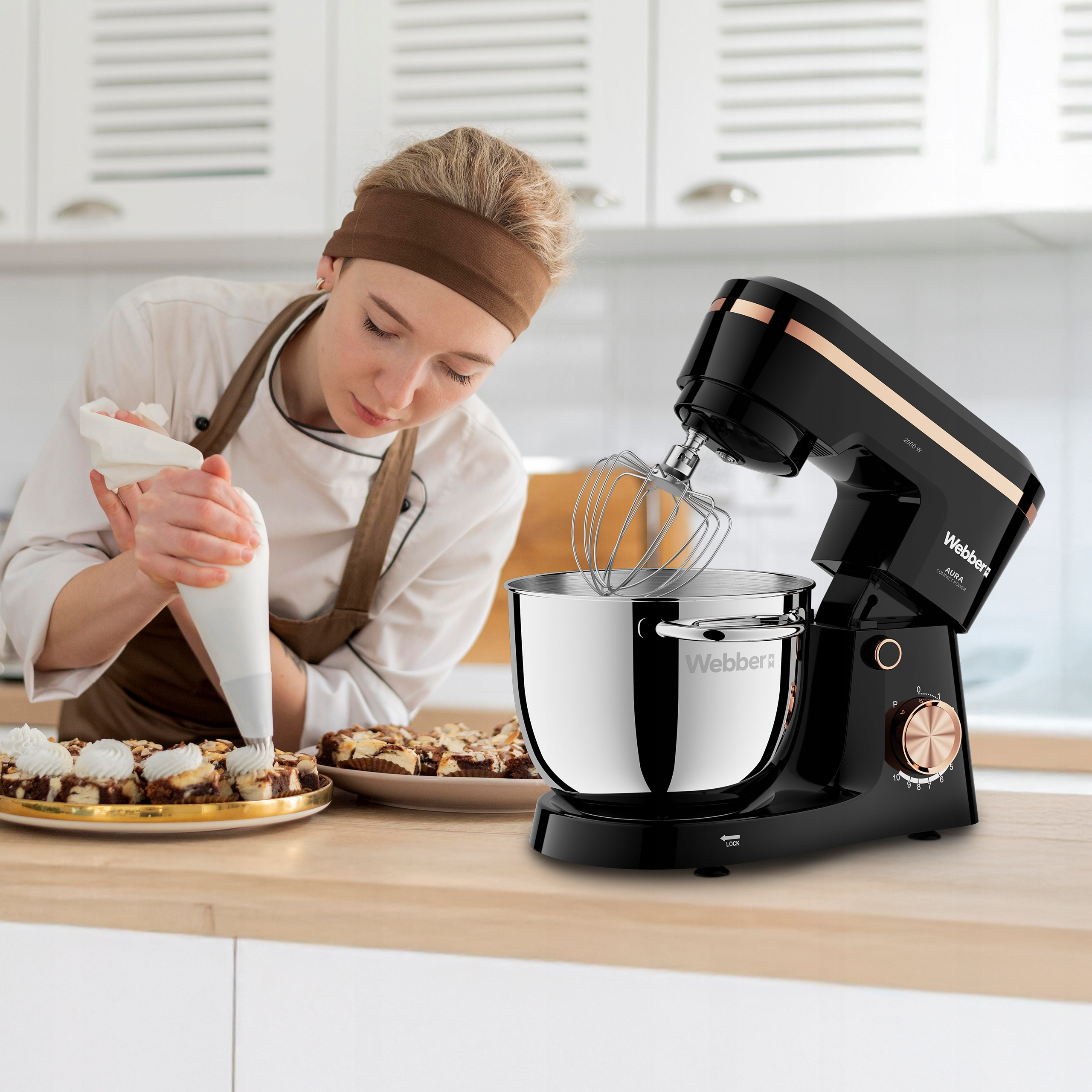 Szef kuchni dekoruje ciasta kremem. Czarny mikser stoi na stole. Ciasta są na tacy.