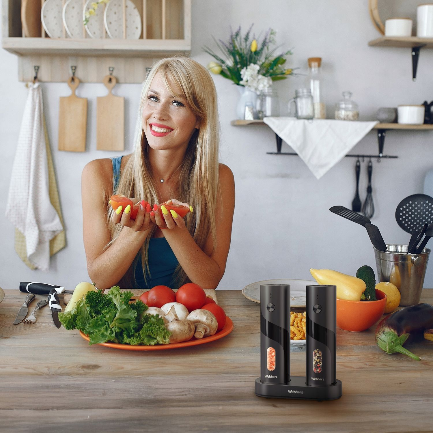 Blondynka w niebieskim topie trzyma pomidory. Talerz warzyw i elektryczne młynki do soli i pieprzu na drewnianym stole.