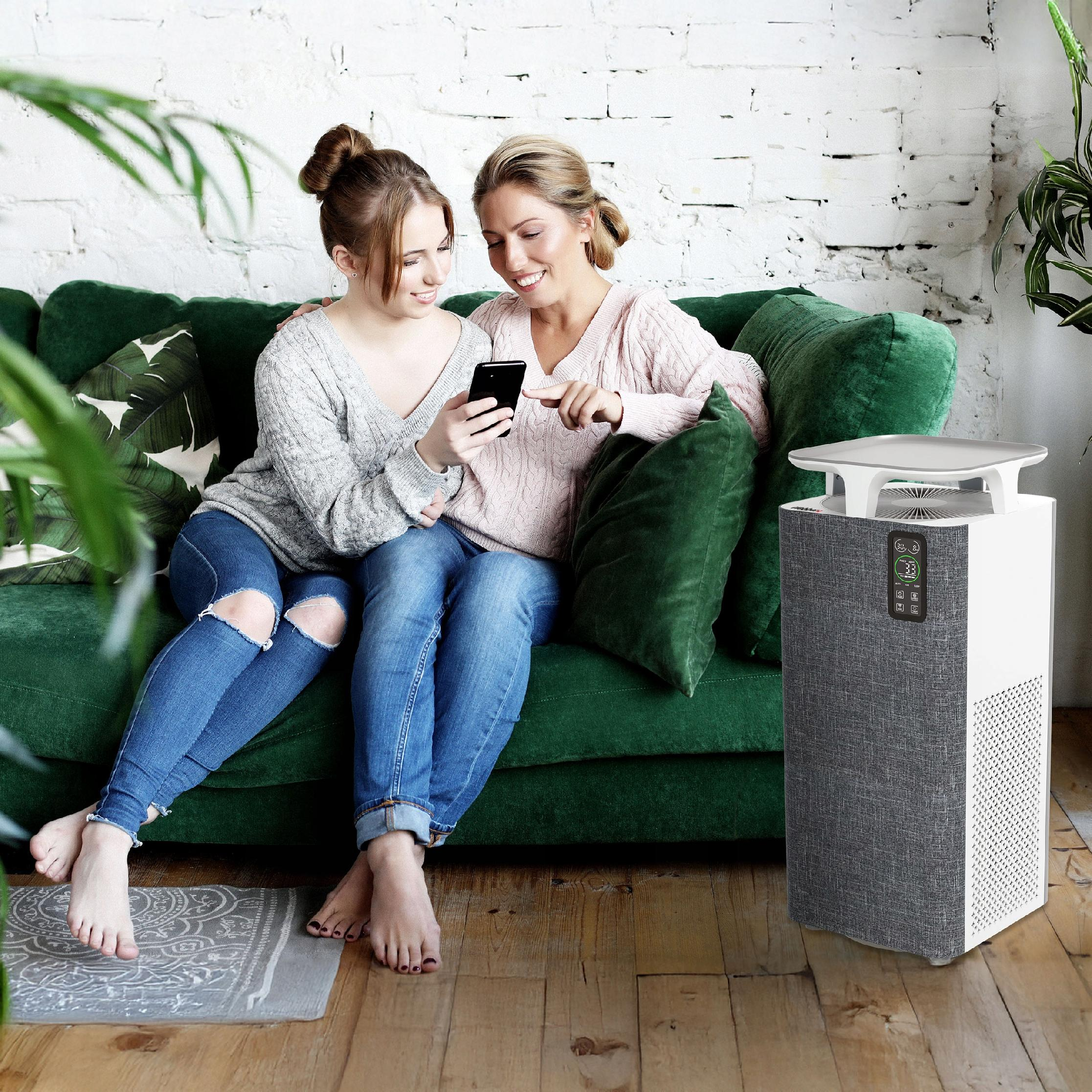 Dwie kobiety na zielonej sofie patrzą na telefon. Oczyszczacz powietrza po prawej. Biała ceglana ściana.
