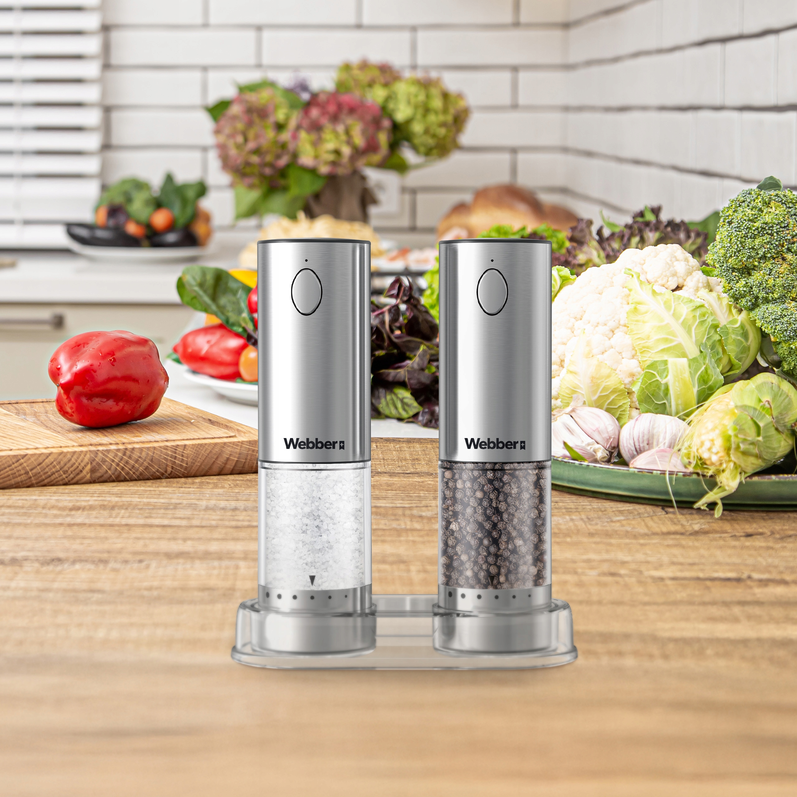 Dwa srebrne młynki do soli i pieprzu Webber na drewnianym stole z warzywami w tle.