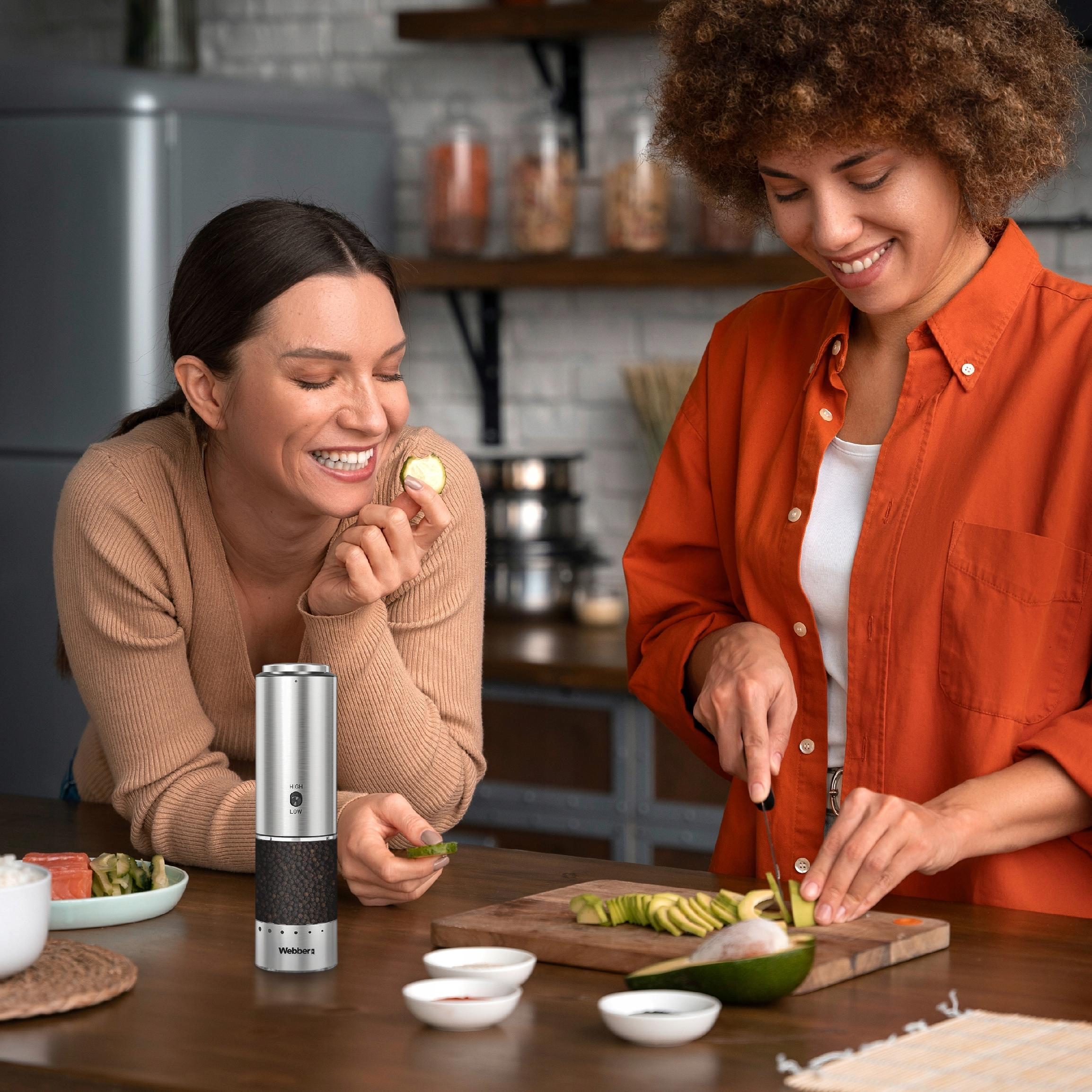 Dwie kobiety w kuchni, jedna kroi awokado. Srebrny młynek i sushi na stole.
