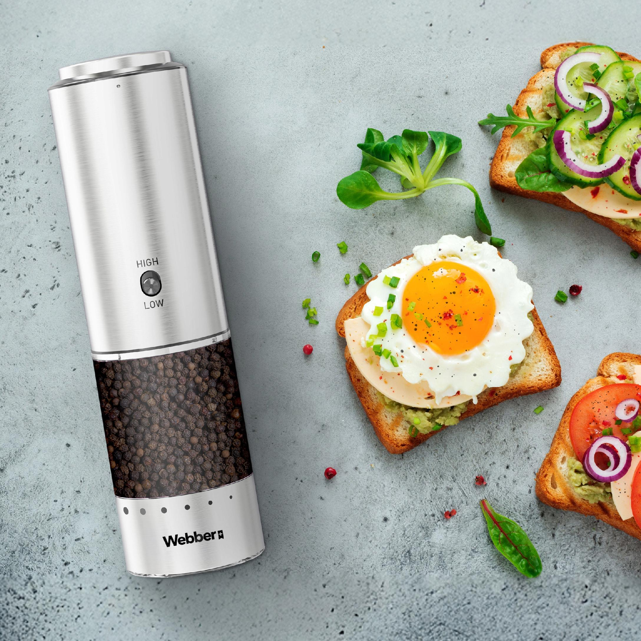 Młynek do pieprzu obok otwartych kanapek z dodatkami, zielenią i jajkiem na szarej powierzchni.