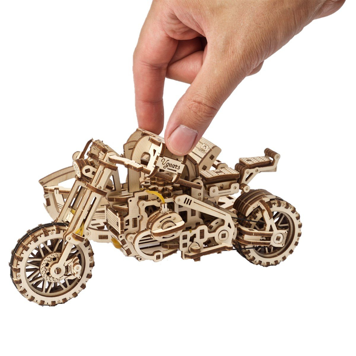 Ręka trzyma drewniany model motocykla. Model wykonany jest z jasnobrązowego drewna, ze szczegółowymi elementami mechanicznymi. Tło jest białe.