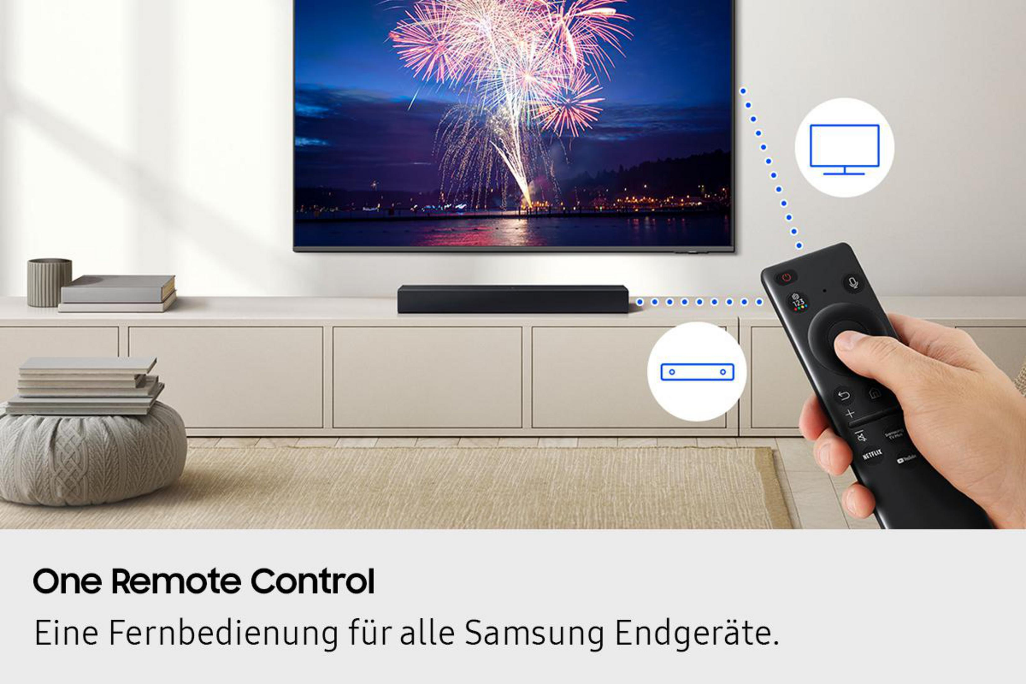 Dłoń trzyma czarny pilot w pobliżu telewizora z fajerwerkami. Pod ekranem znajduje się soundbar.