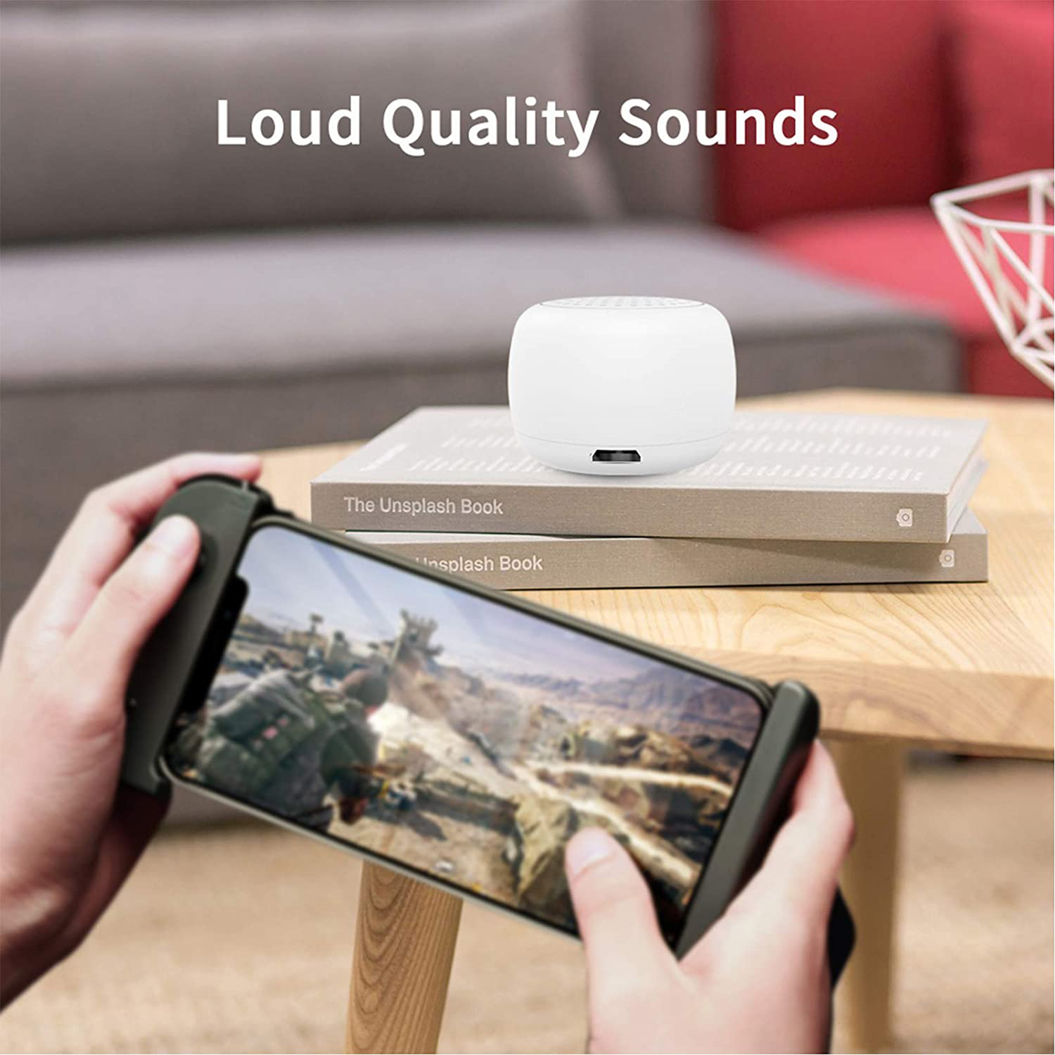 Ein weißer Lautsprecher liegt auf Büchern. Ein Telefon mit einem Spiel wird gehalten. Im Hintergrund sieht man ein Sofa und Text: Loud Quality Sounds.