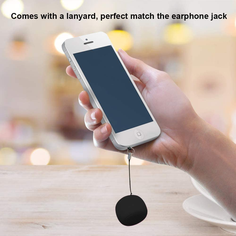 Hand hält ein weißes Smartphone mit einem an einen schwarzen, runden Lautsprecher befestigten Lanyard. Der Hintergrund zeigt eine Holzoberfläche.