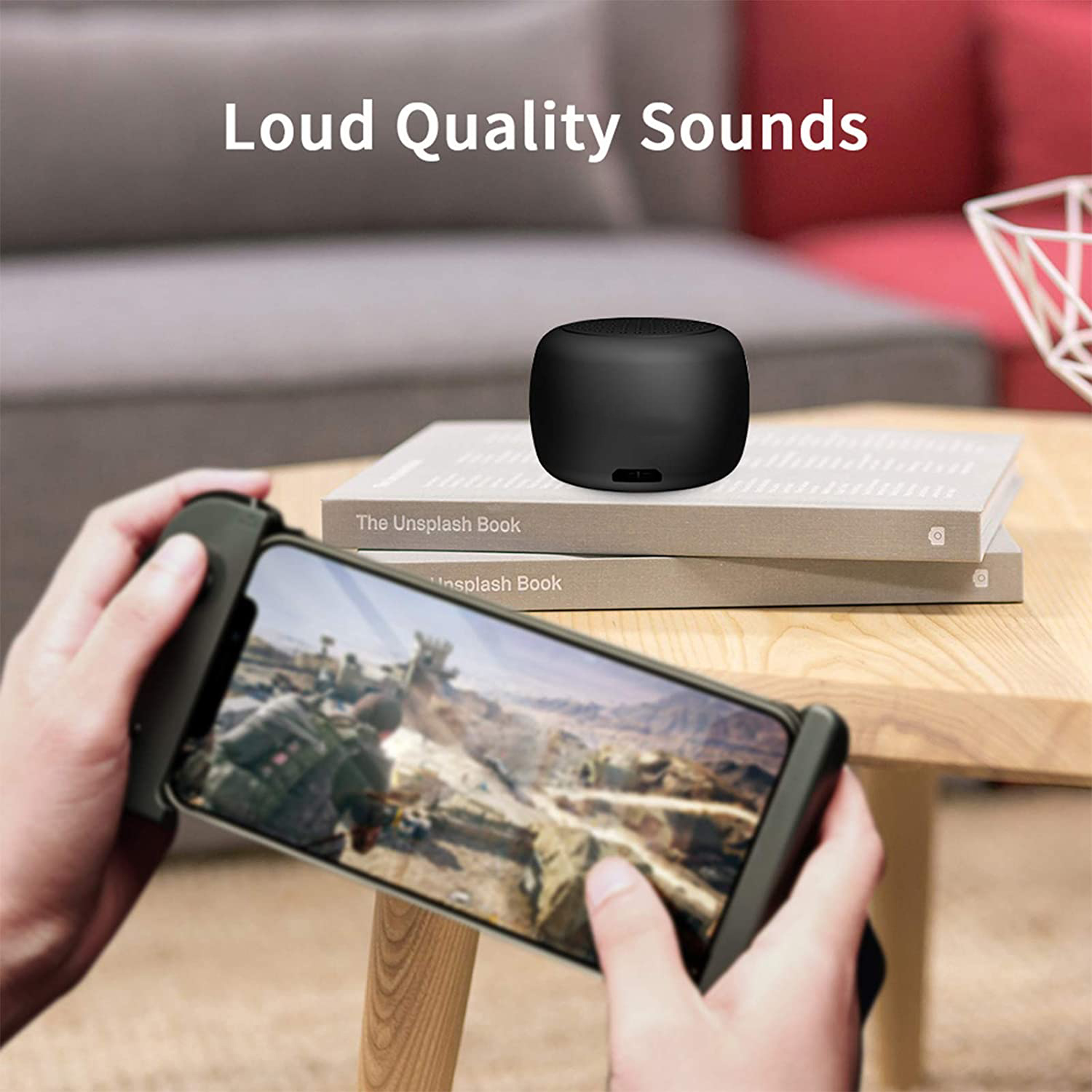 Ein schwarzer Lautsprecher liegt auf Büchern. Ein Telefon mit Spiel wird gehalten. Text lautet: 'Loud Quality Sounds'.