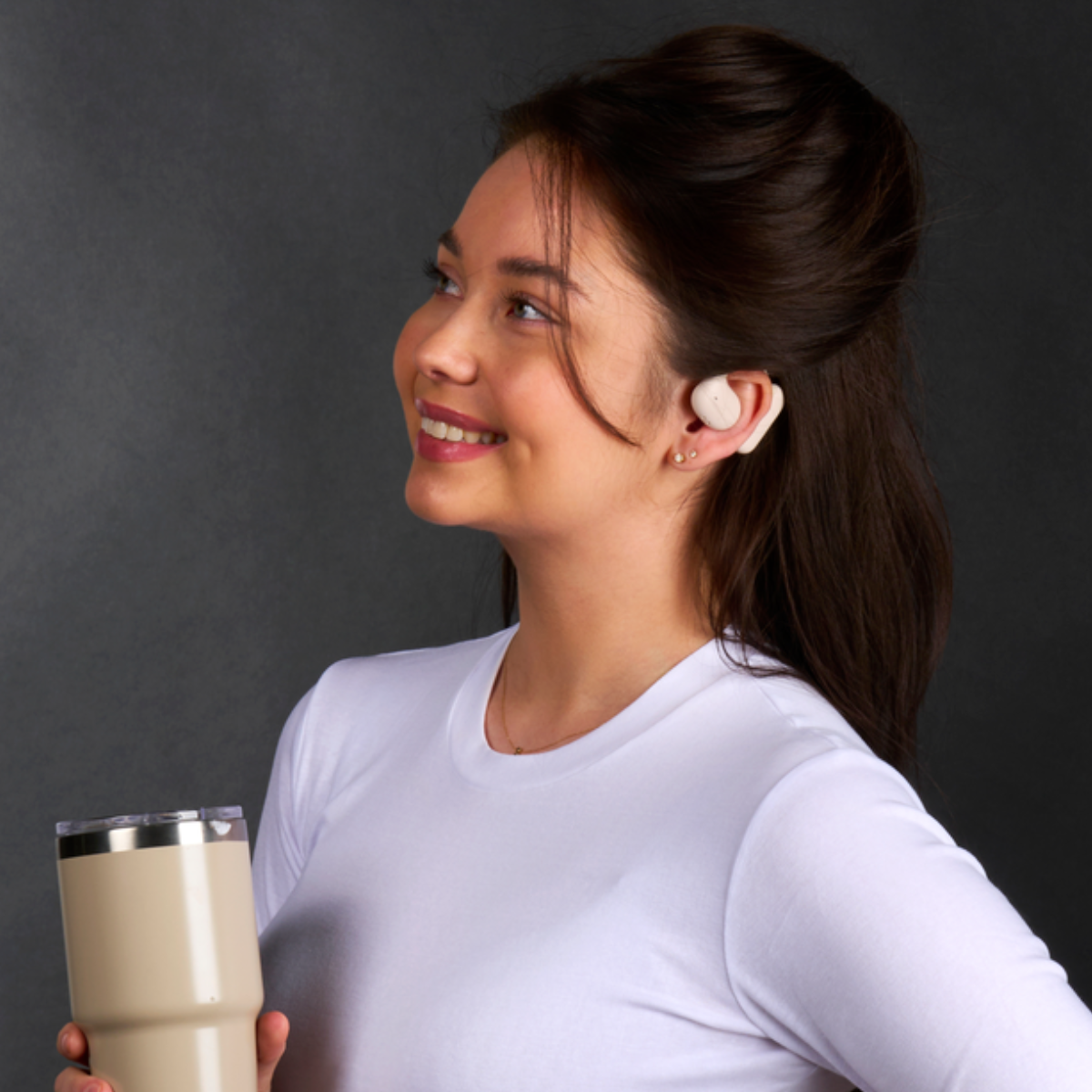 Smiling woman in white shirt with earbuds, holding a beige tumbler. Dark background.