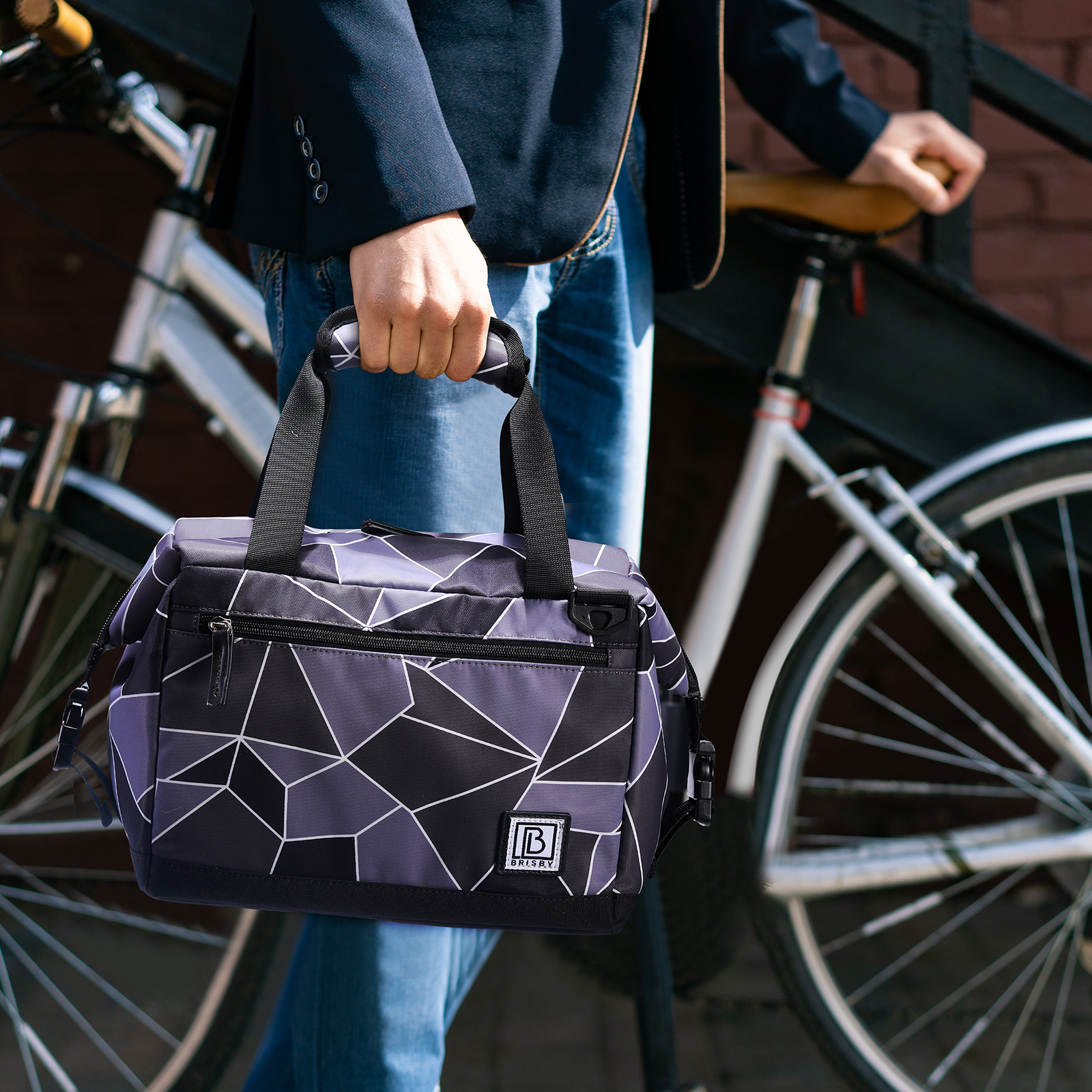 Een persoon houdt een tas met geometrisch patroon vast. Een fiets staat achter hen. De persoon draagt een blazer en jeans. Een bakstenen muur is zichtbaar.