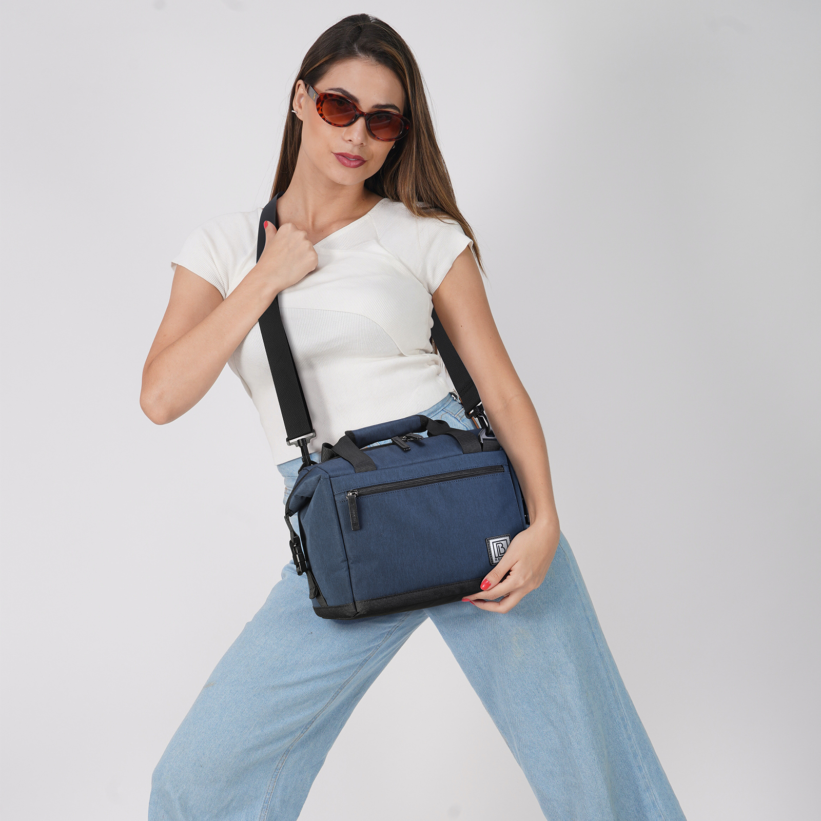 Vrouw met zonnebril, witte top en jeans houdt een blauwe tas met zwarte riem vast. Neutrale achtergrond.