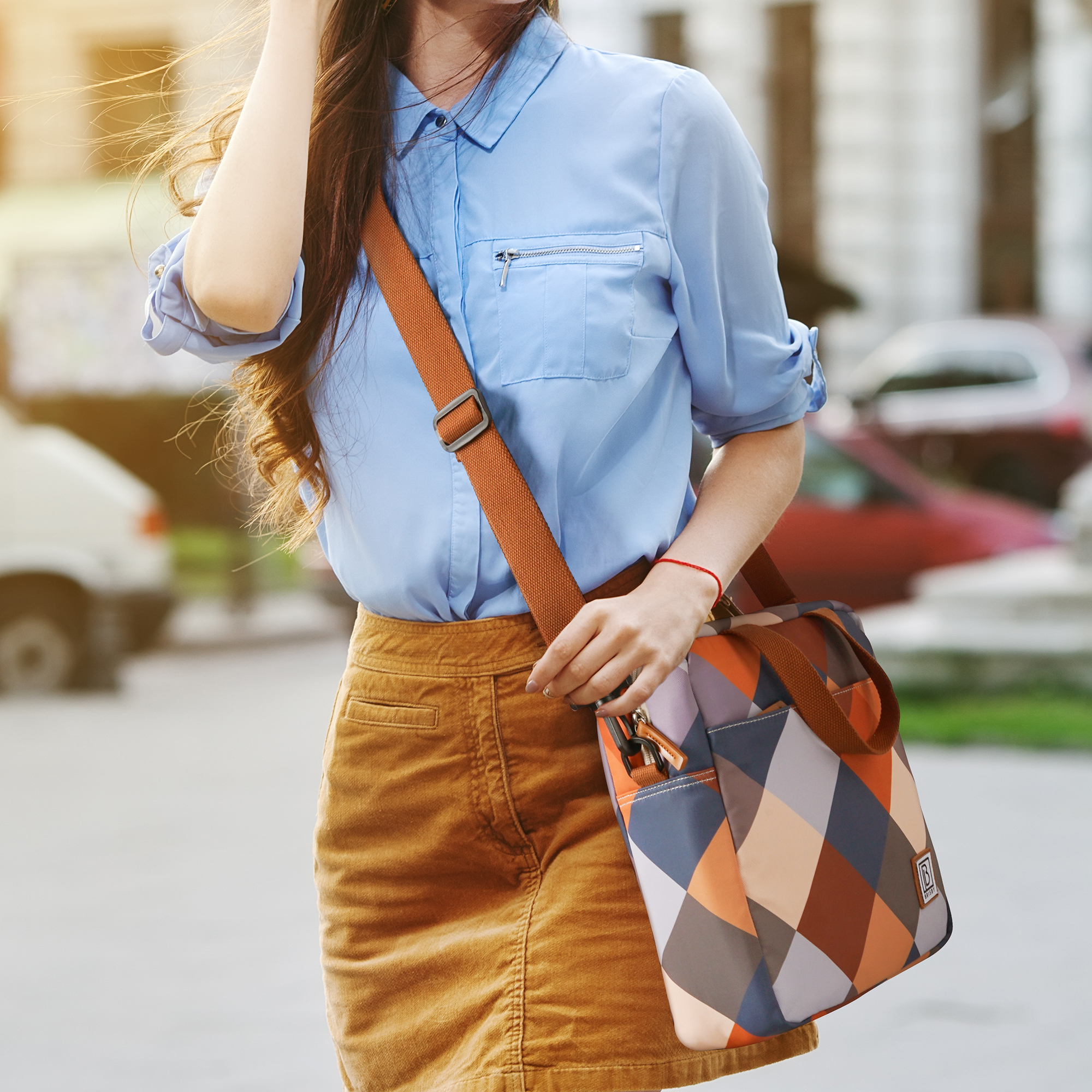Vrouw in blauw overhemd, bruine rok en tas met patroon, lopend. Stad achtergrond met auto's.