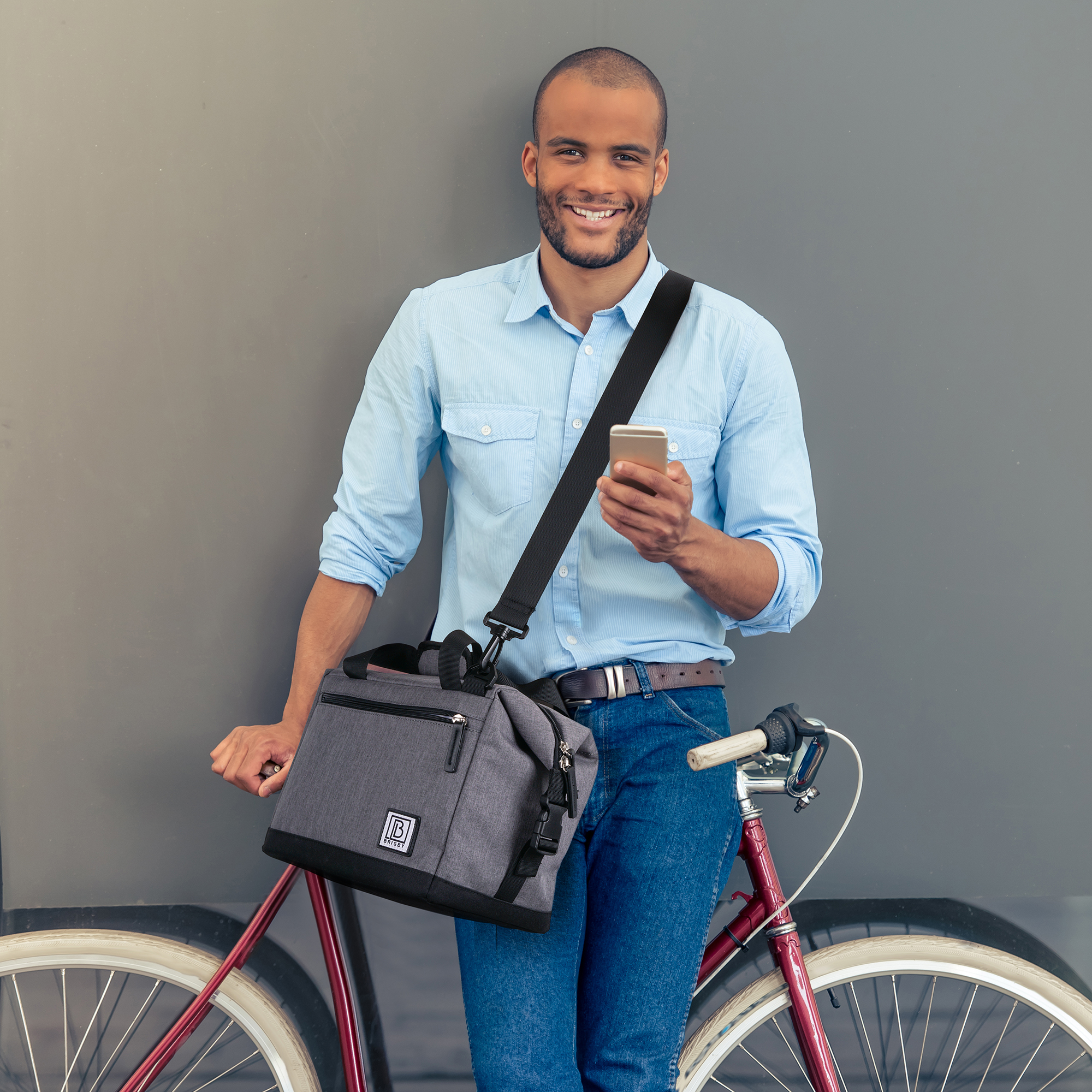 Een man houdt een telefoon vast, leunt op een fiets en draagt een shirt en jeans. Hij heeft een tas over zijn schouder.