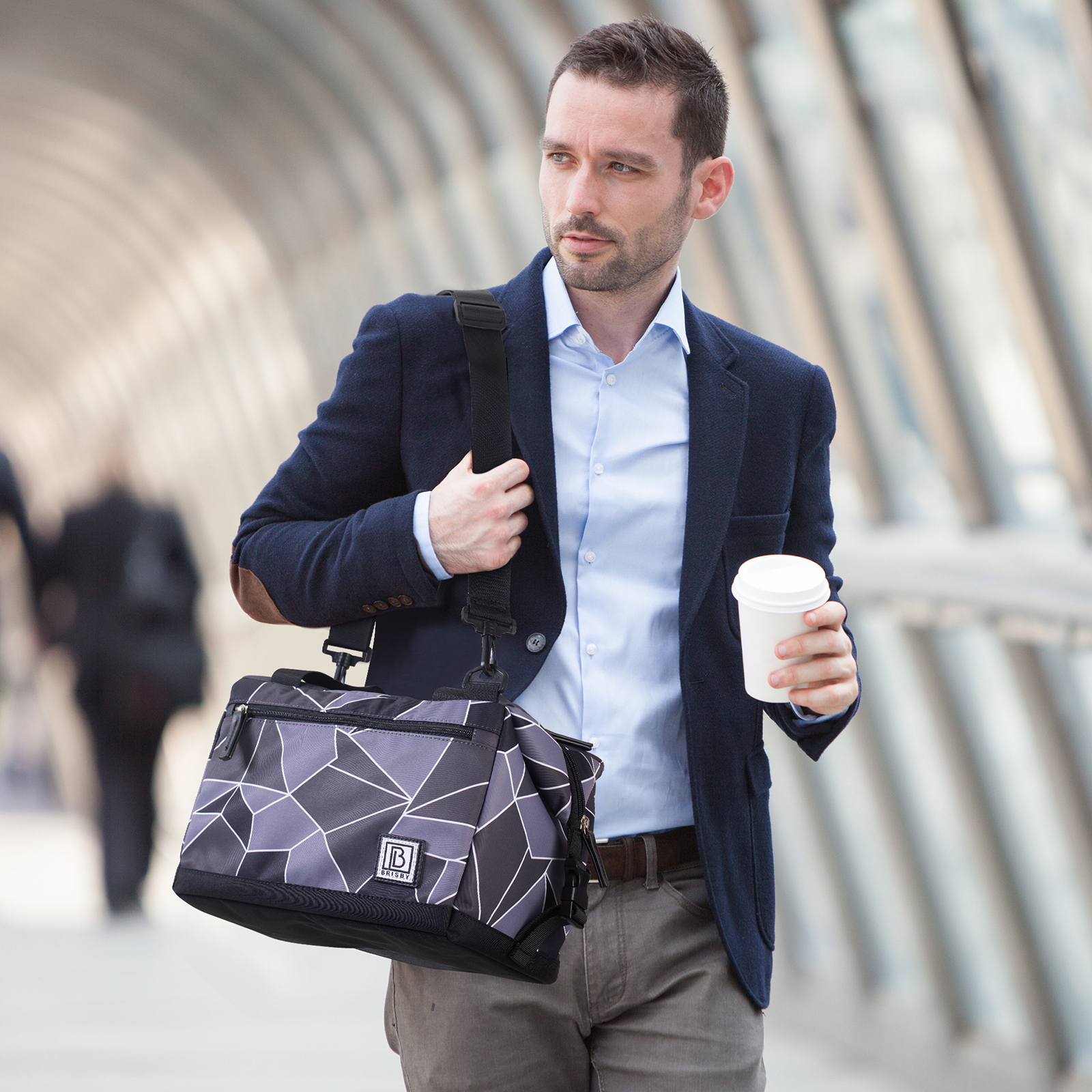 Man in pak loopt, draagt een tas en koffie. Achtergrond is een moderne loopbrug. Hij kijkt naar rechts.